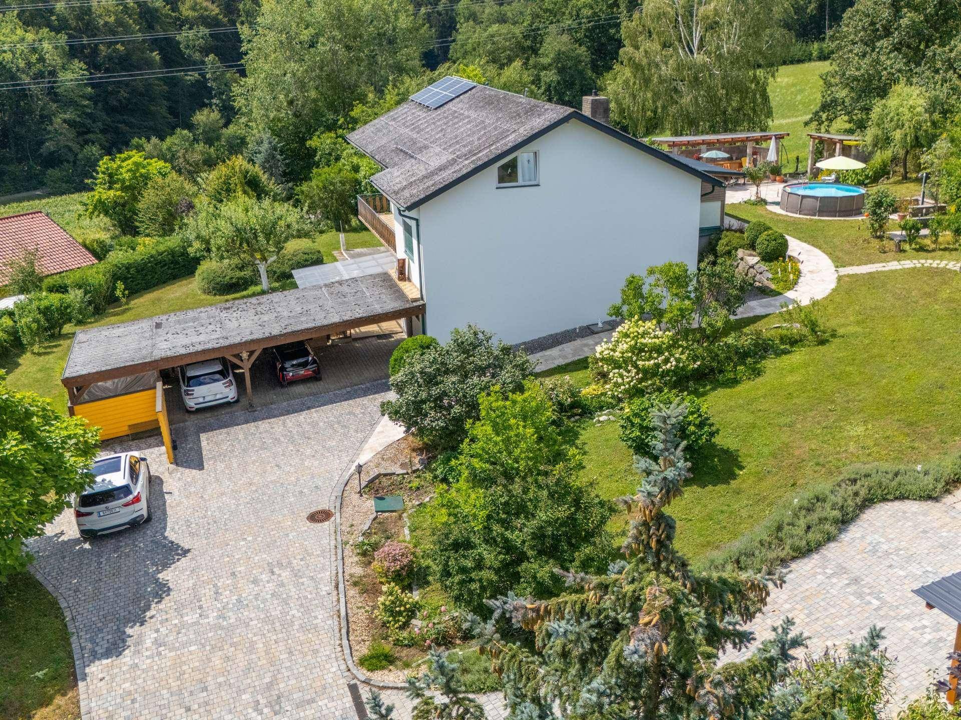 Luftaufnahme des Hauses mit Carport, Gartenanlage und einem Pool im Hintergrund.