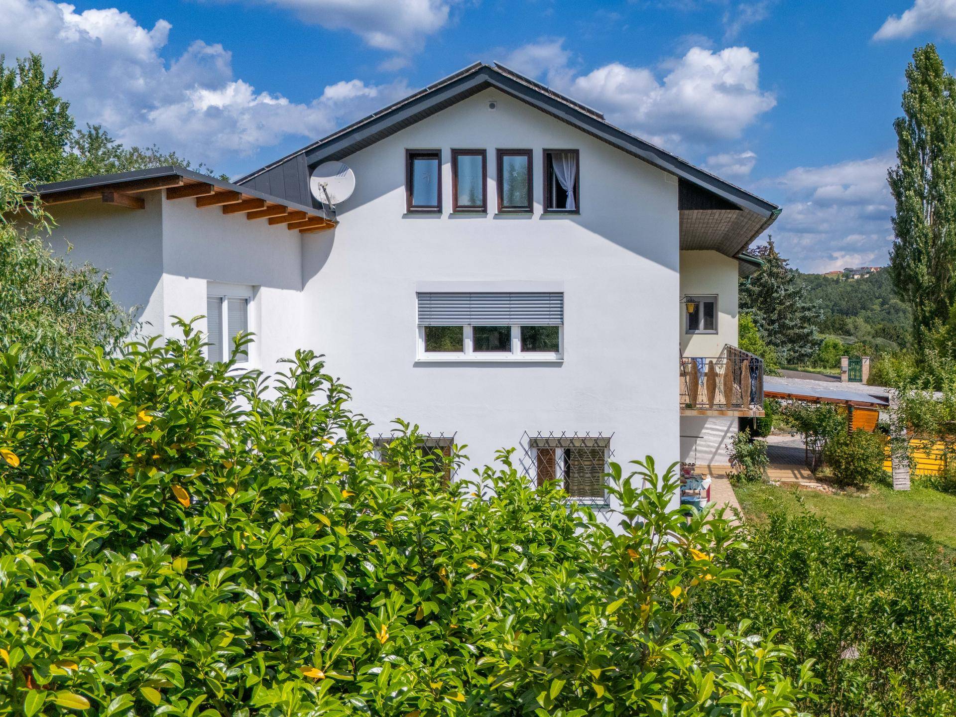 Weiße Hausfassade mit mehreren Fenstern und einem Balkon, umgeben von grünen Büschen.