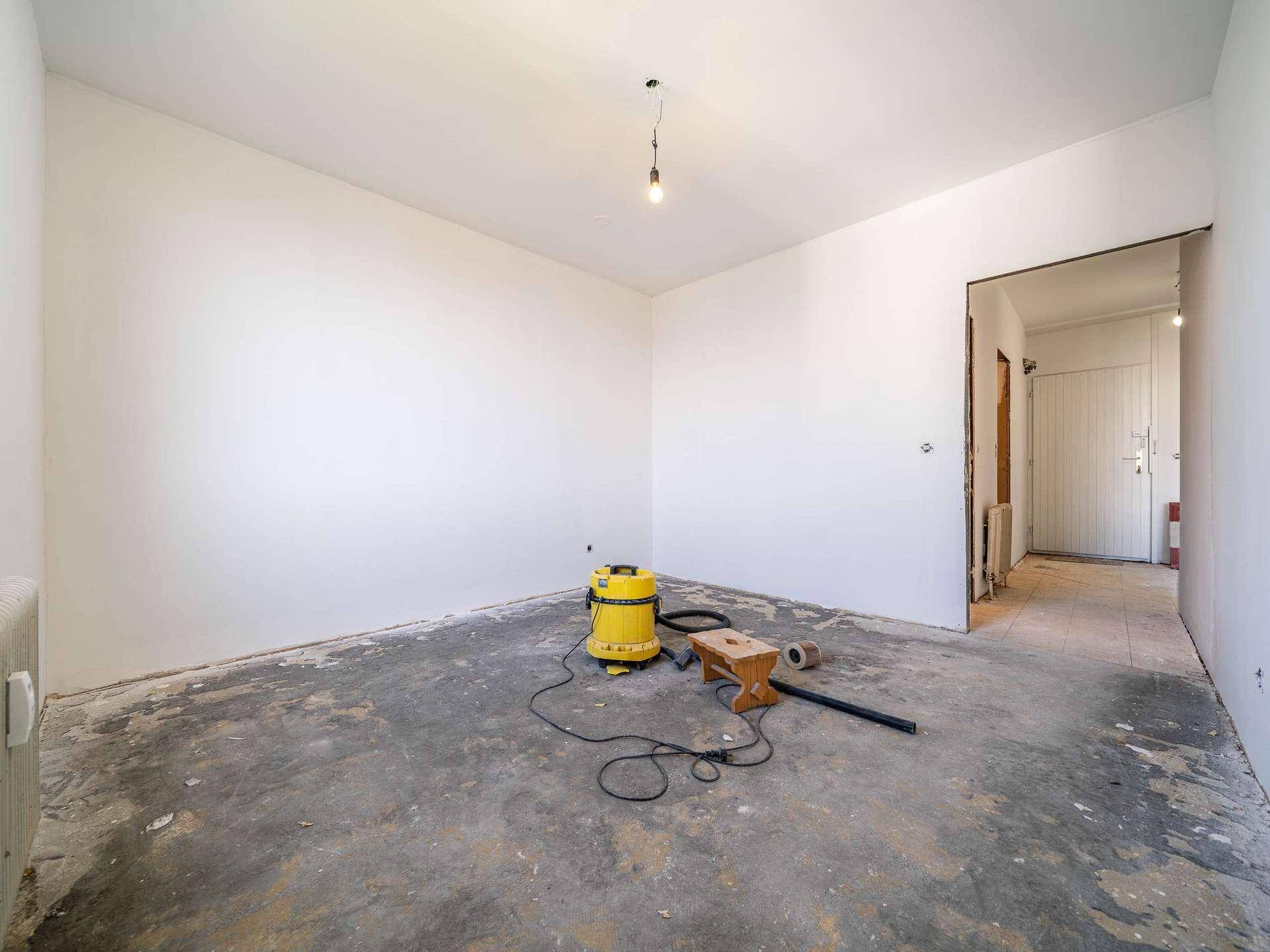 Renovierungsbedürftiges Zimmer mit Reinigungsgeräten und Blick in den Flur der Wohnung.
