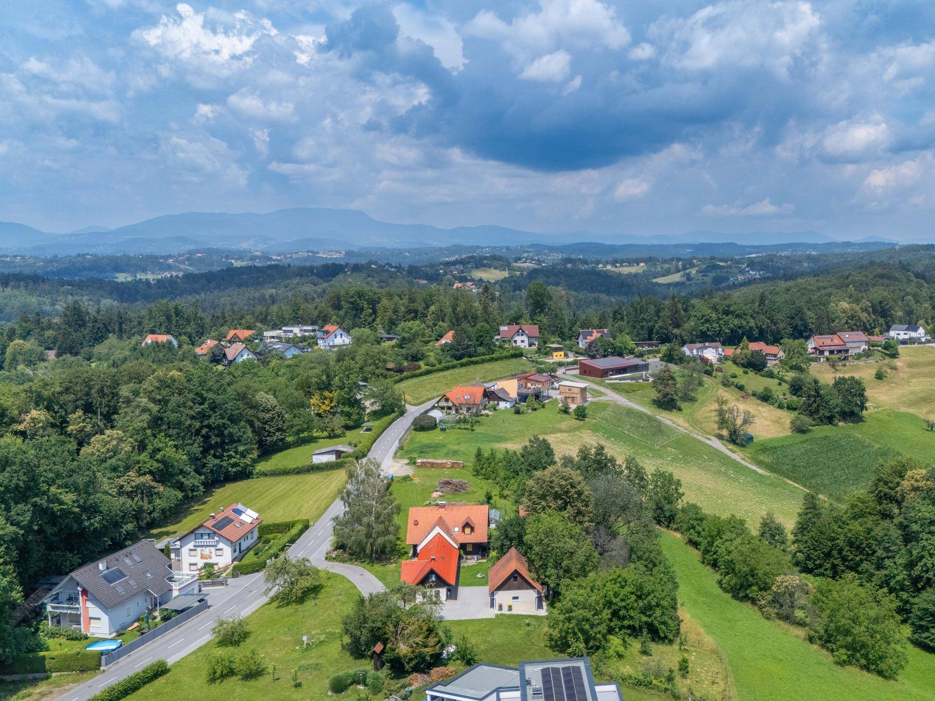 Luftaufnahme einer ländlichen Siedlung in einem Tal, umgeben von Wäldern und Feldern mit Bergblick.