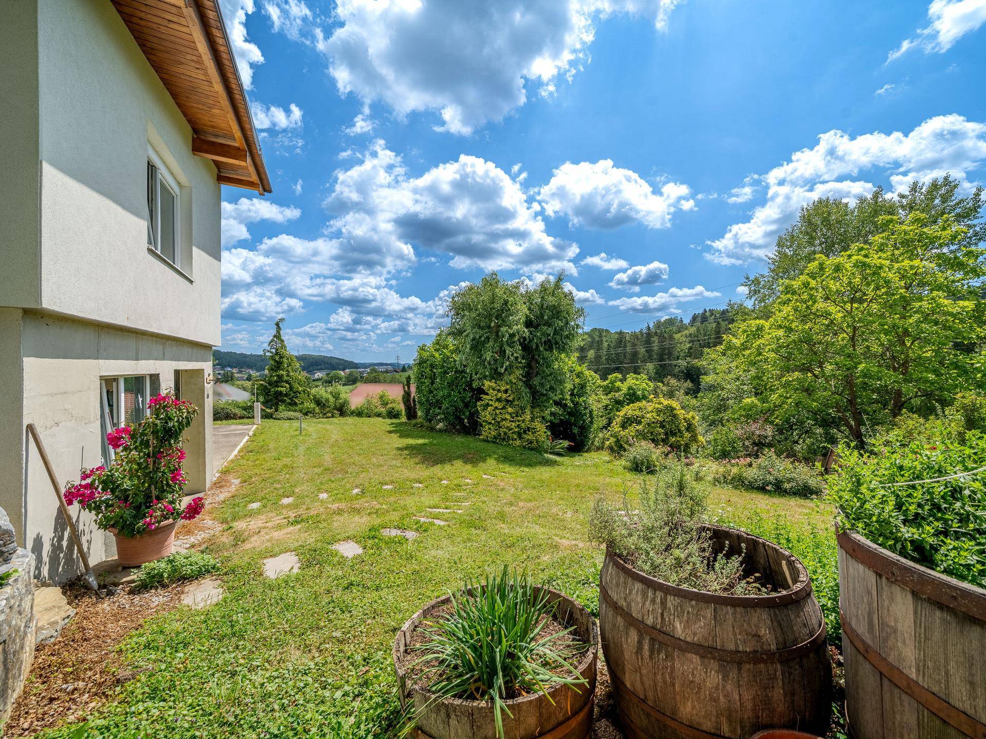 Hausfassade mit gepflegtem Gartenbereich und dekorativen Holzfässern unter blauem Himmel.