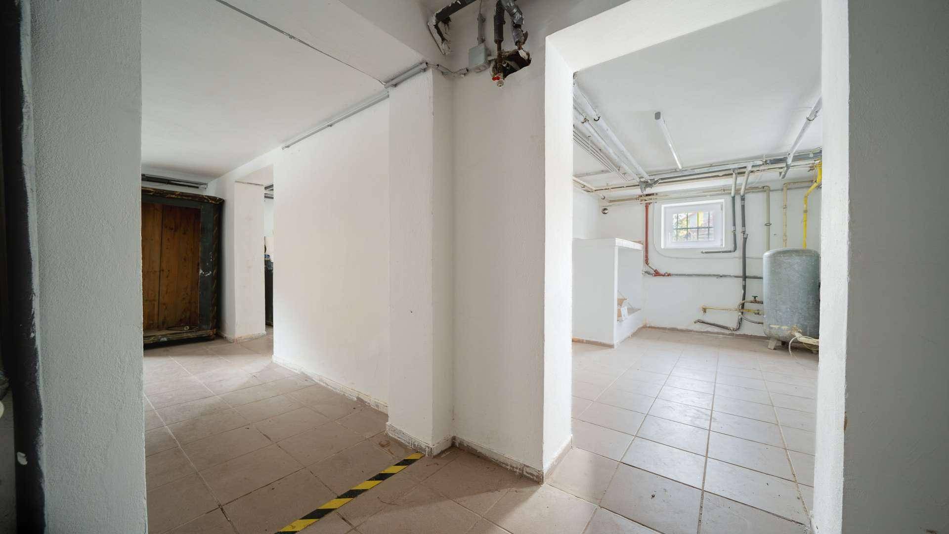 Kellerraum mit Fliesenboden, sichtbaren Rohren und Zugang zu weiteren Kellerräumen.