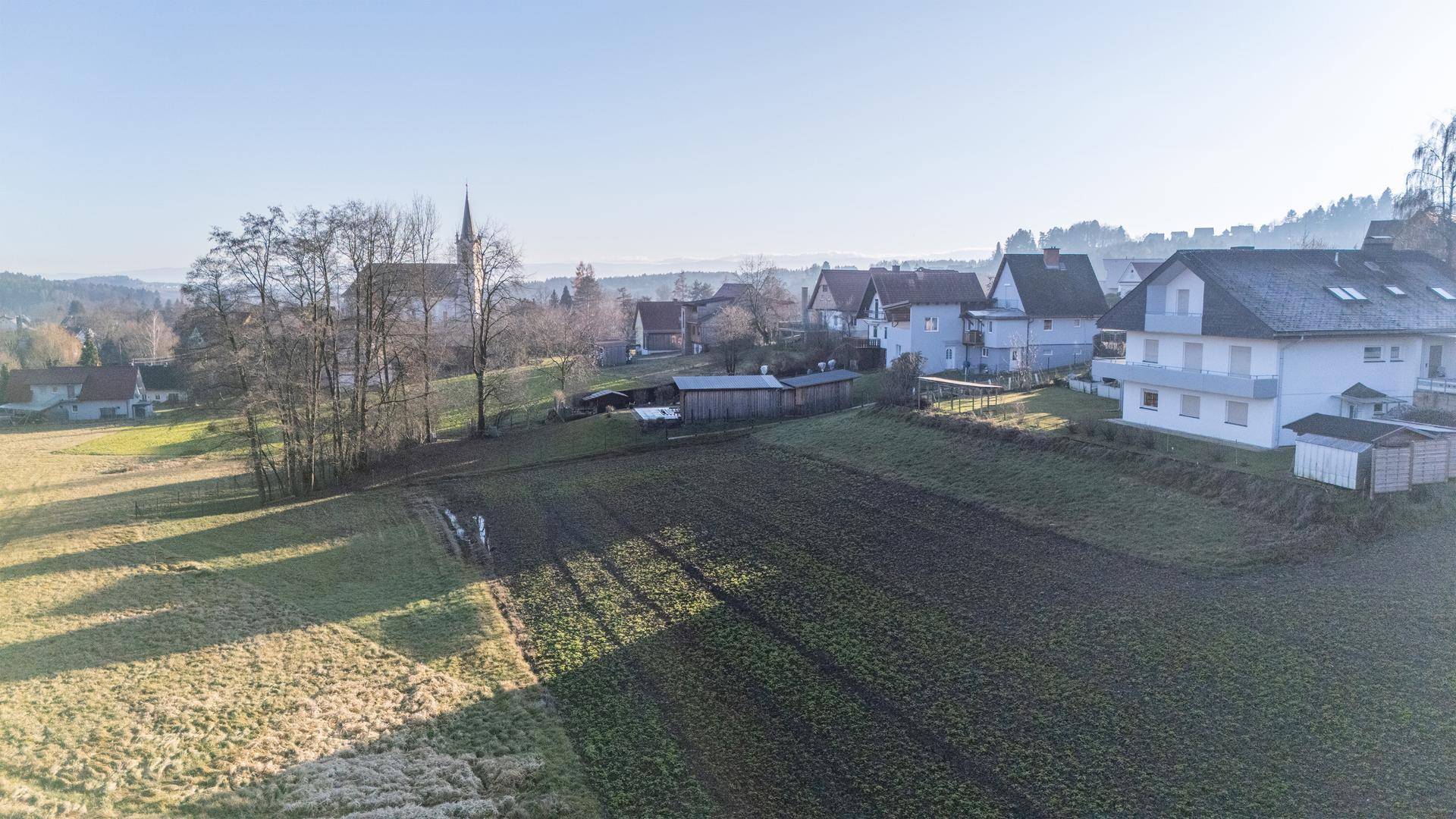 Blick über eine ländliche Siedlung mit Wohnhäusern, Feldern und der Dorfkirche im Hintergrund an einem sonnigen Tag.