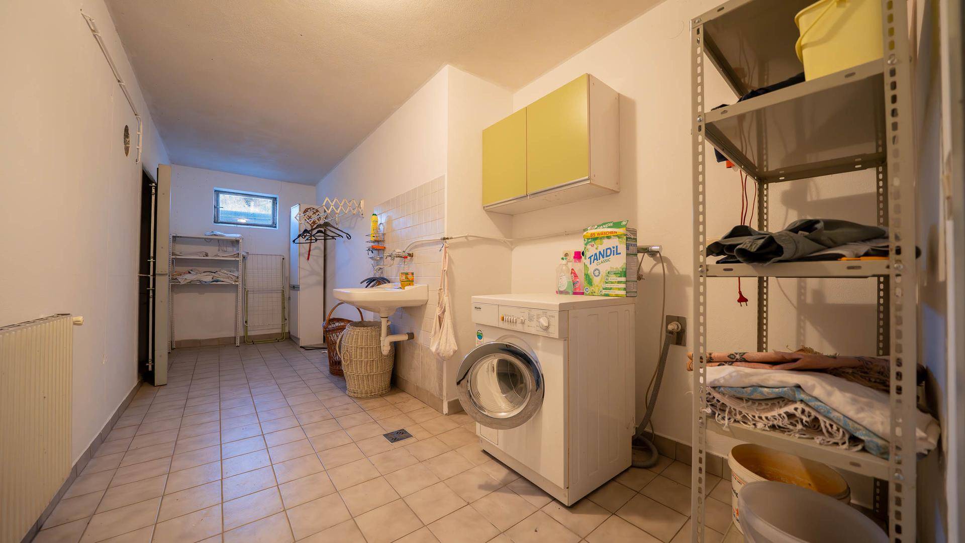 Waschküche im Keller mit Waschmaschine, Spülbecken und Regalen.