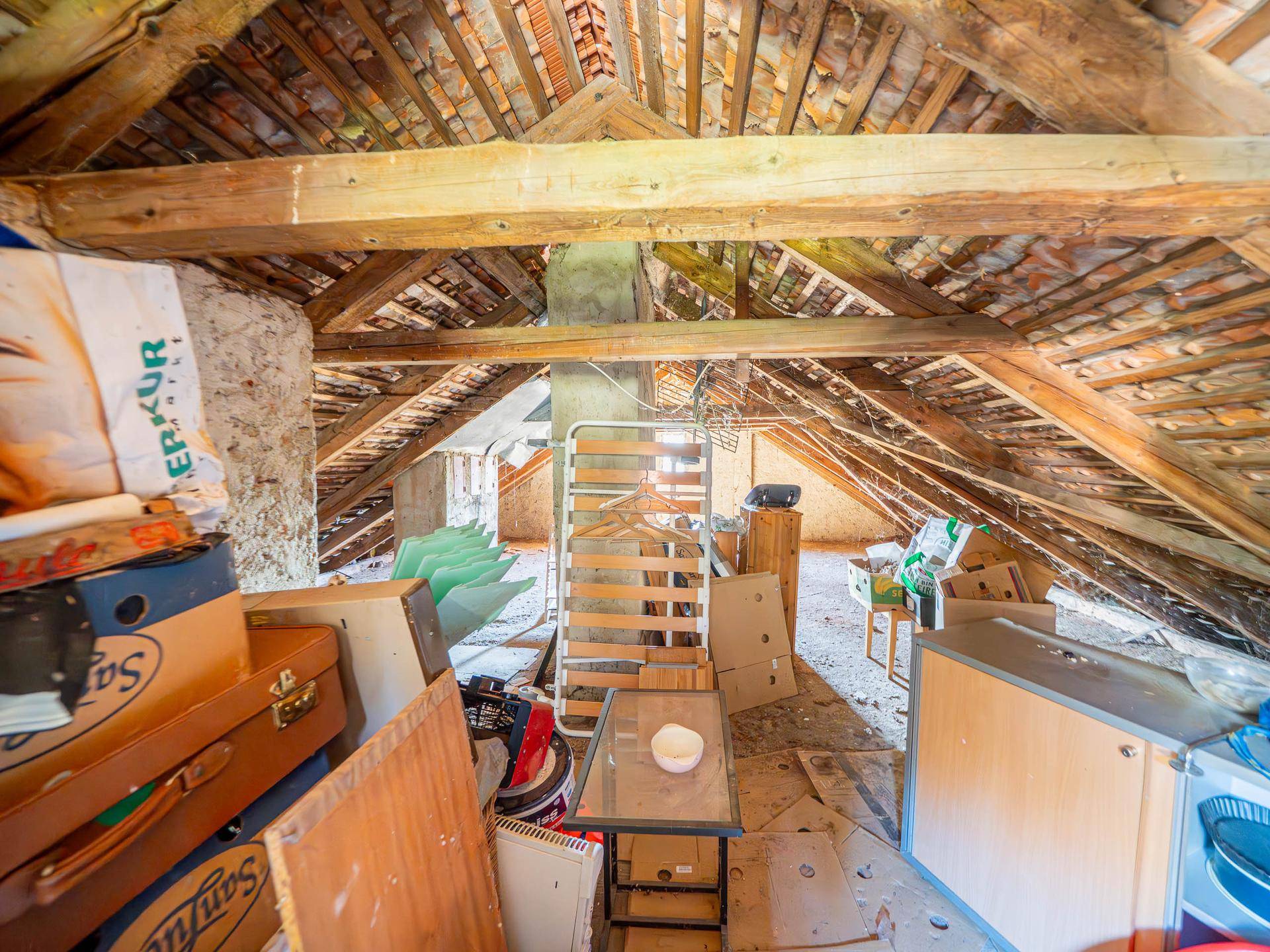 Unausgebauter Dachboden mit sichtbaren Holzbalken und Ziegeln, der als Lagerraum dient.