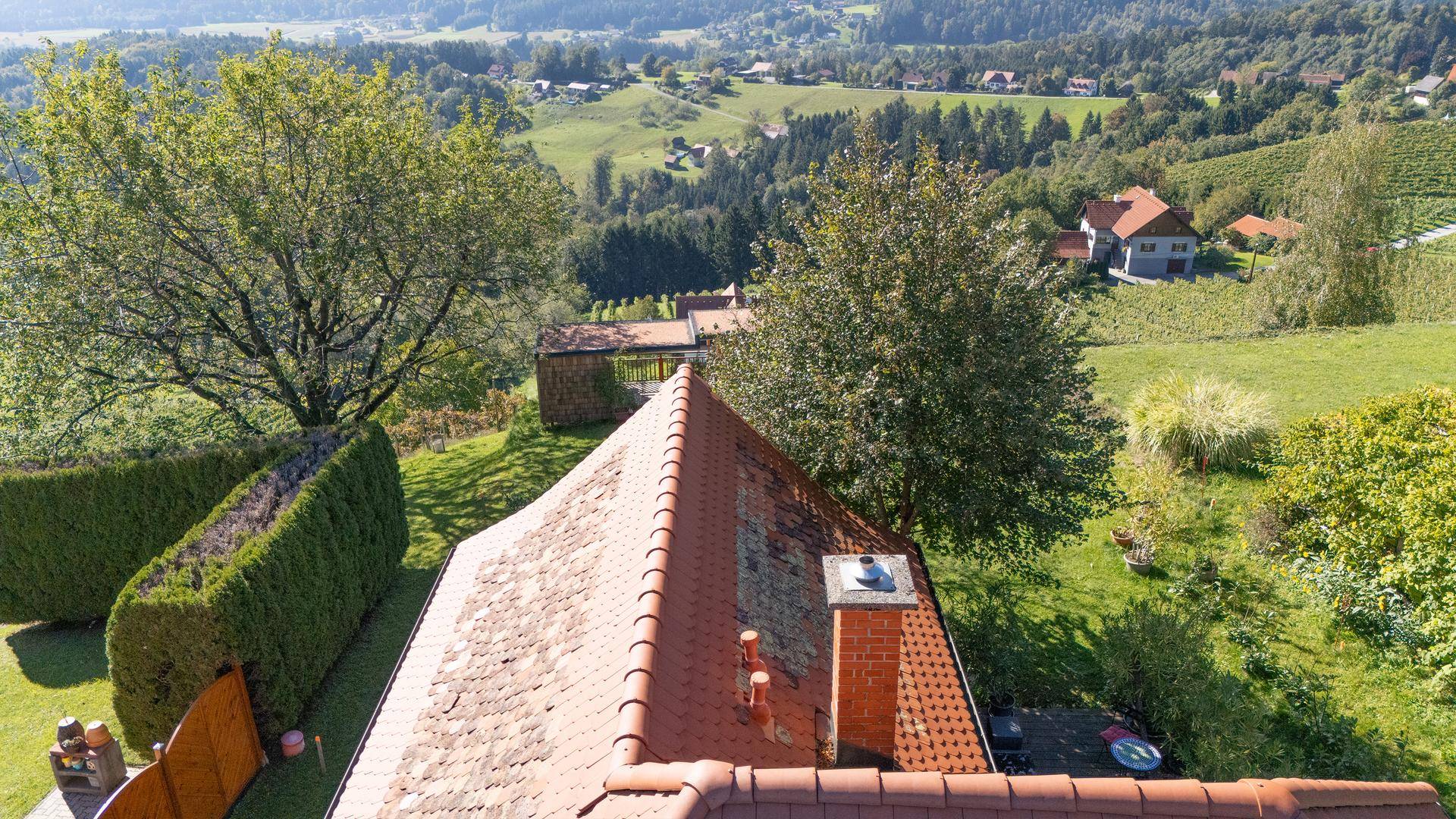 Ausblick über ein Ziegeldach mit Kamin auf Bäume und eine sanfte Hügellandschaft.