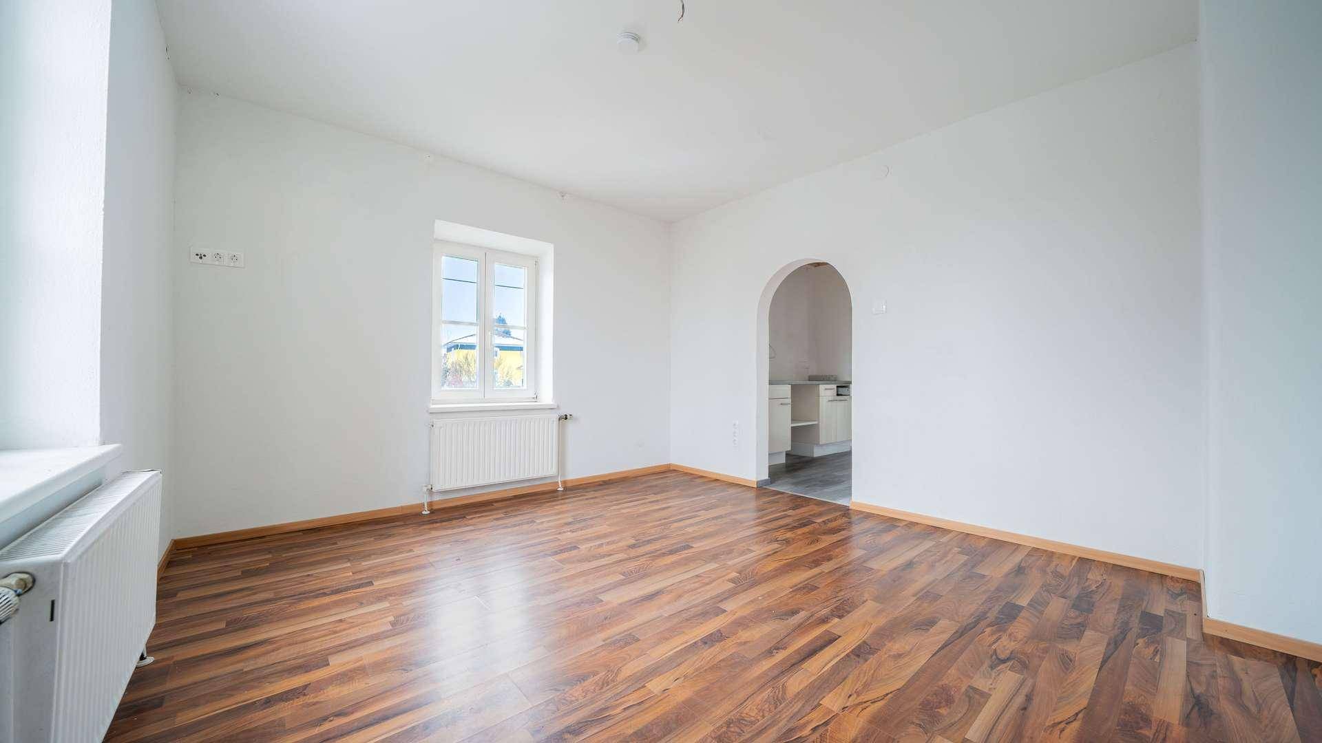 Geräumiges Wohnzimmer mit dunklem Laminatboden, weiß gestrichenen Wänden und einem Fenster.