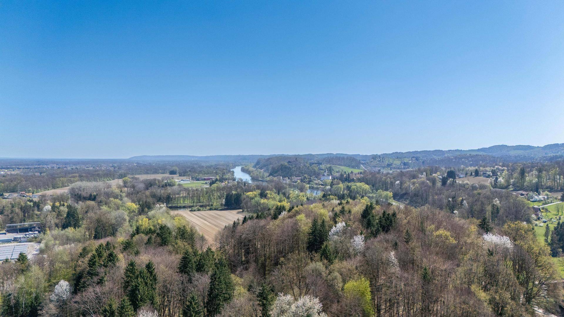 Panoramablick über eine weite, grüne Landschaft mit Wäldern, Feldern und einem Fluss in der Ferne.