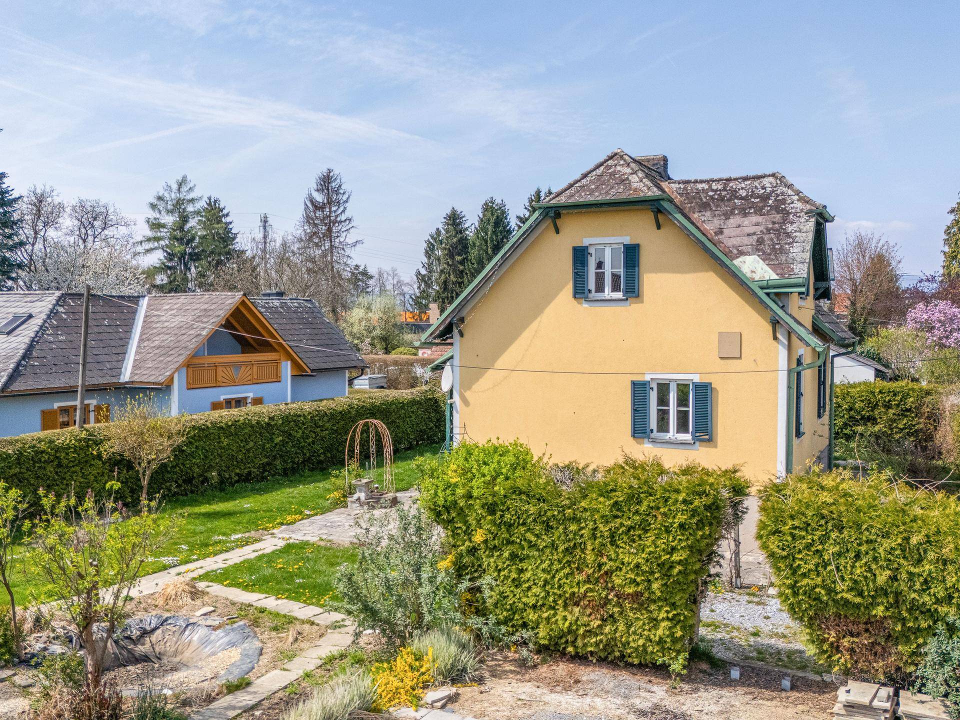 Seitenansicht des Hauses mit gelber Fassade, umgeben von einem grünen Garten mit Hecken und Bäumen.
