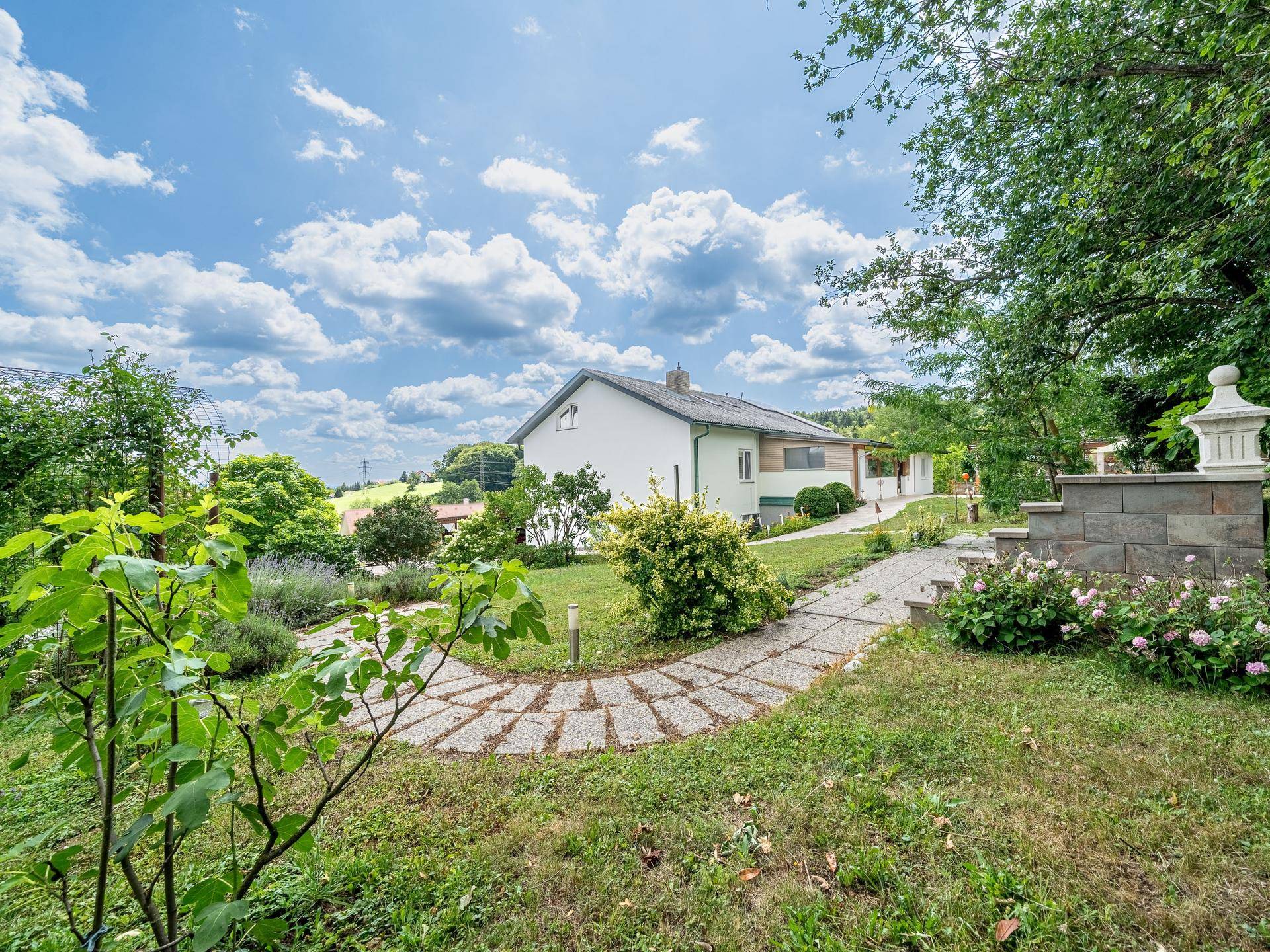 Geschwungener Gartenweg durch eine grüne Anlage mit Blumenbeeten und Blick auf das Haus.
