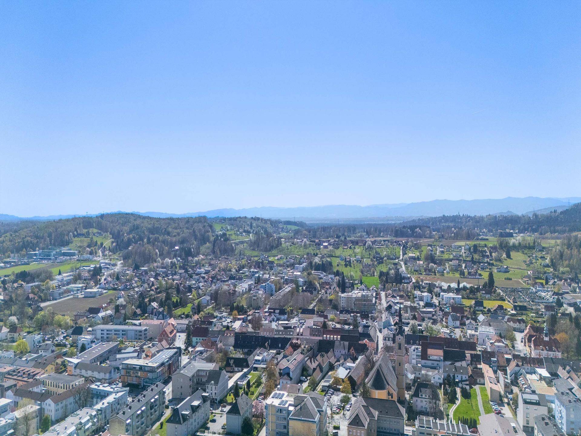 Weitläufige Luftaufnahme einer Stadt mit zahlreichen Gebäuden und sanften Hügeln im Hintergrund.