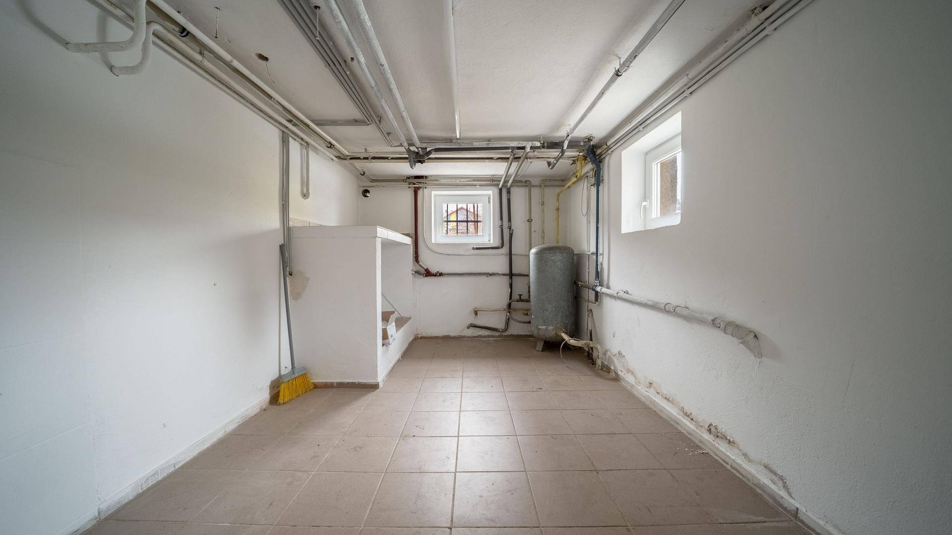 Funktionaler Kellerraum mit Fliesenboden, sichtbaren Installationen und Fenstern, dient als Technikraum.