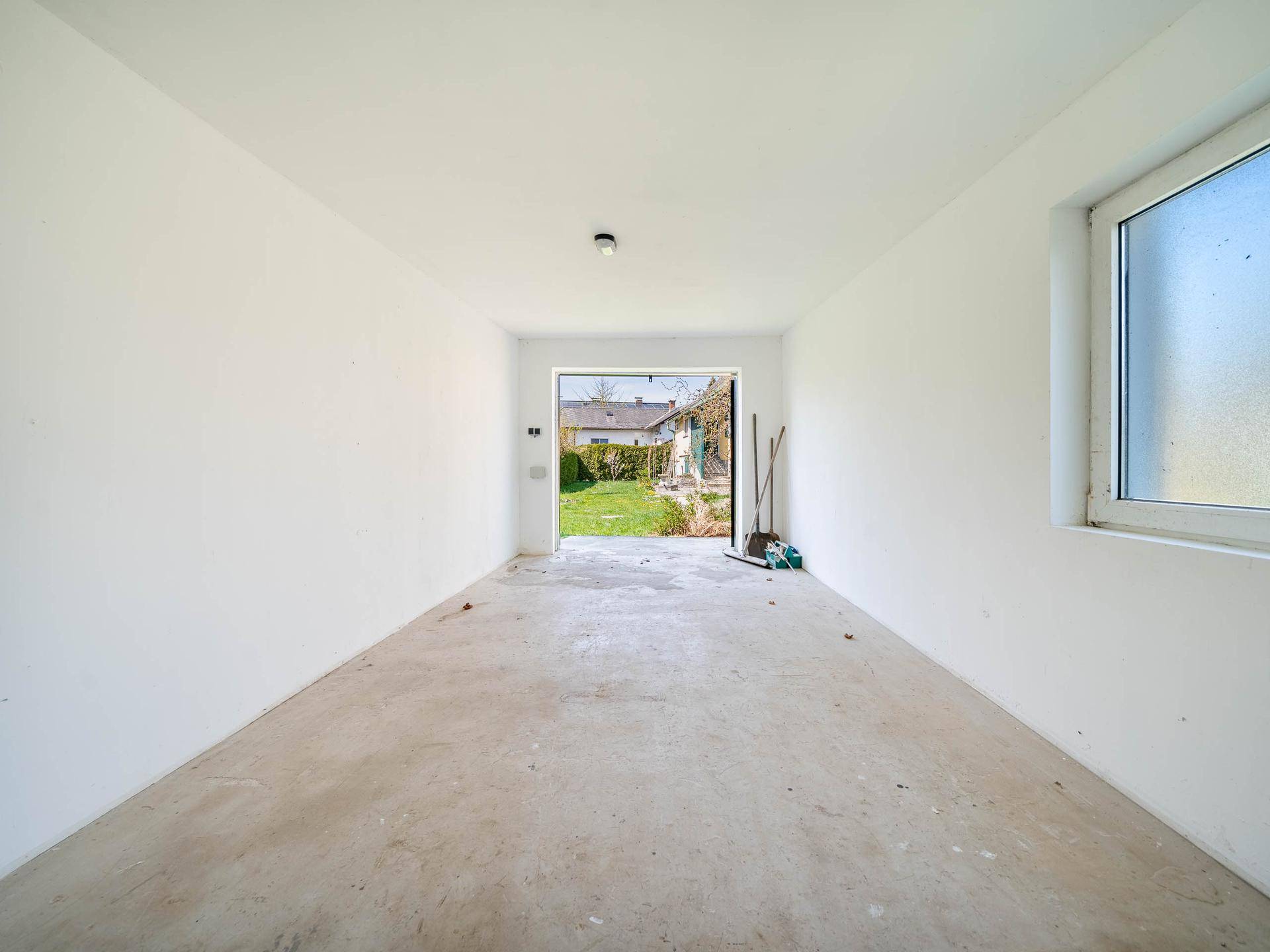 Leerer Innenraum des Nebengebäudes mit Betonboden und einem Fenster, ideal als Garage oder Lagerraum.