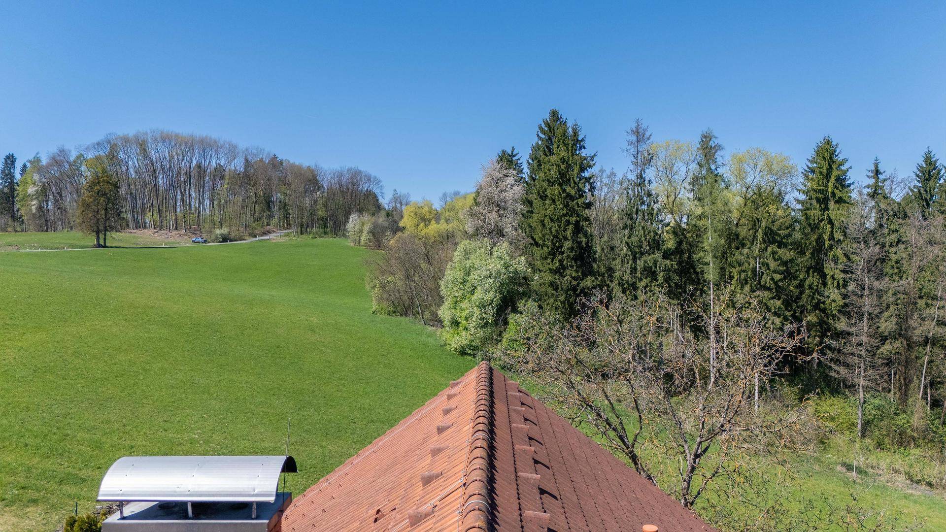 Blick über das Ziegeldach des Hauses auf weite grüne Wiesen und dichte Wälder.