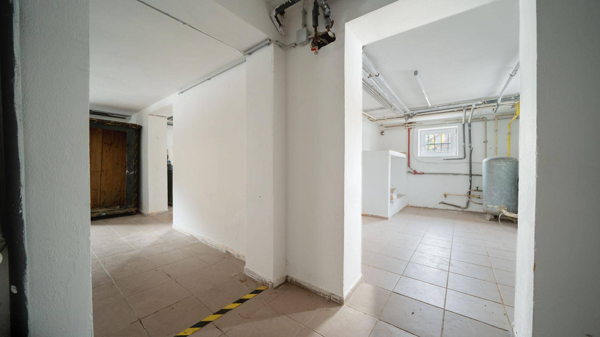 Kellerbereich mit Fliesenboden, sichtbaren Leitungen und einem Fenster, ideal als Abstellraum.