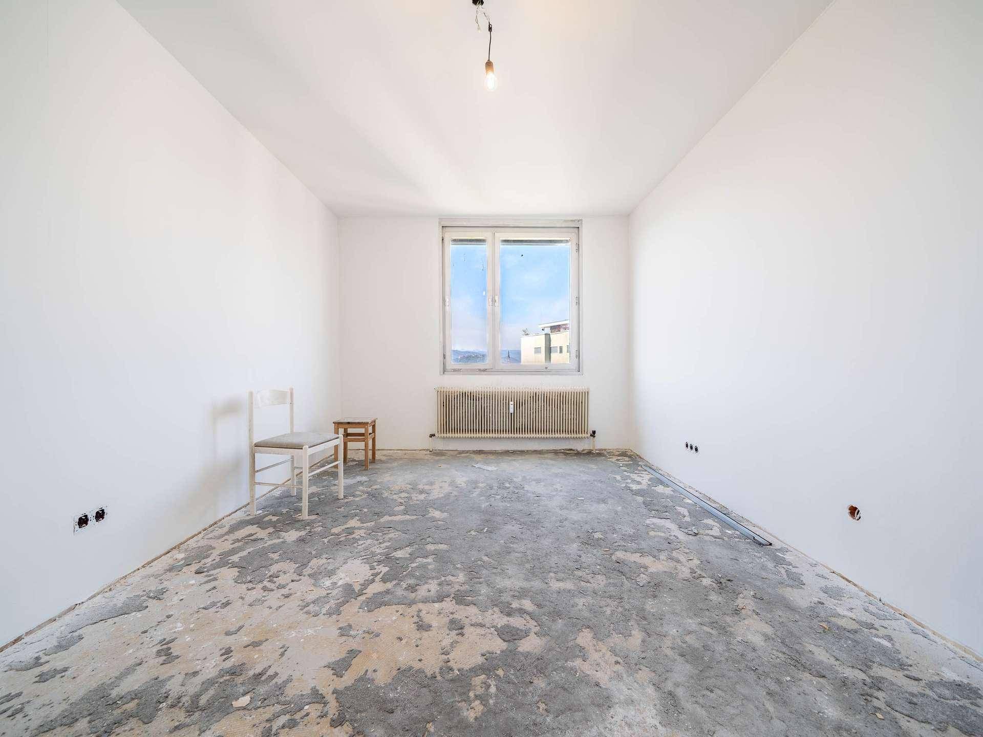 Leeres Zimmer in Renovierung mit großem Fenster, Heizkörper und abgenutztem Bodenbelag.