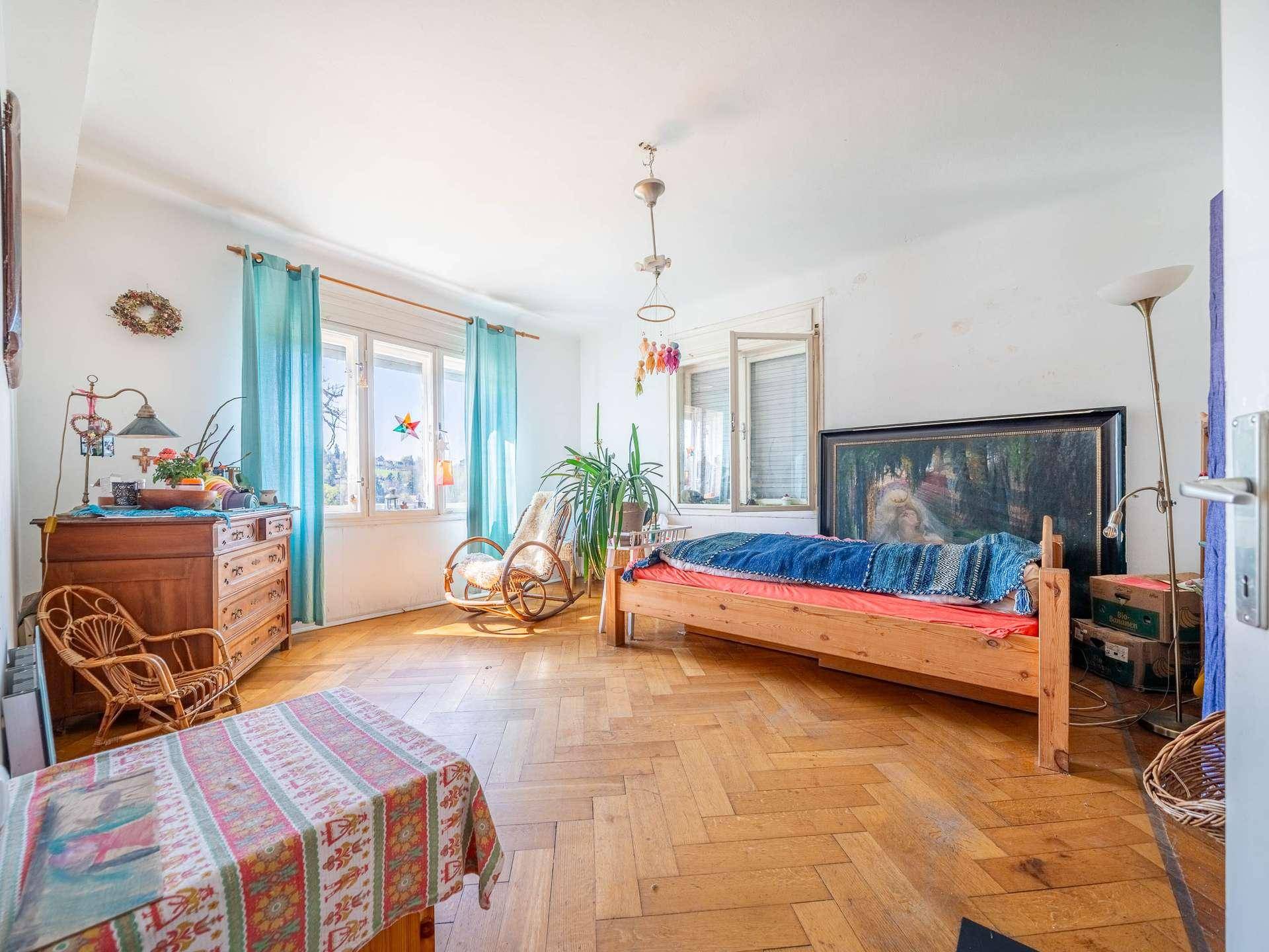 Helles Schlafzimmer mit Parkettboden, Holzmöbeln und großen Fenstern mit blauen Vorhängen.