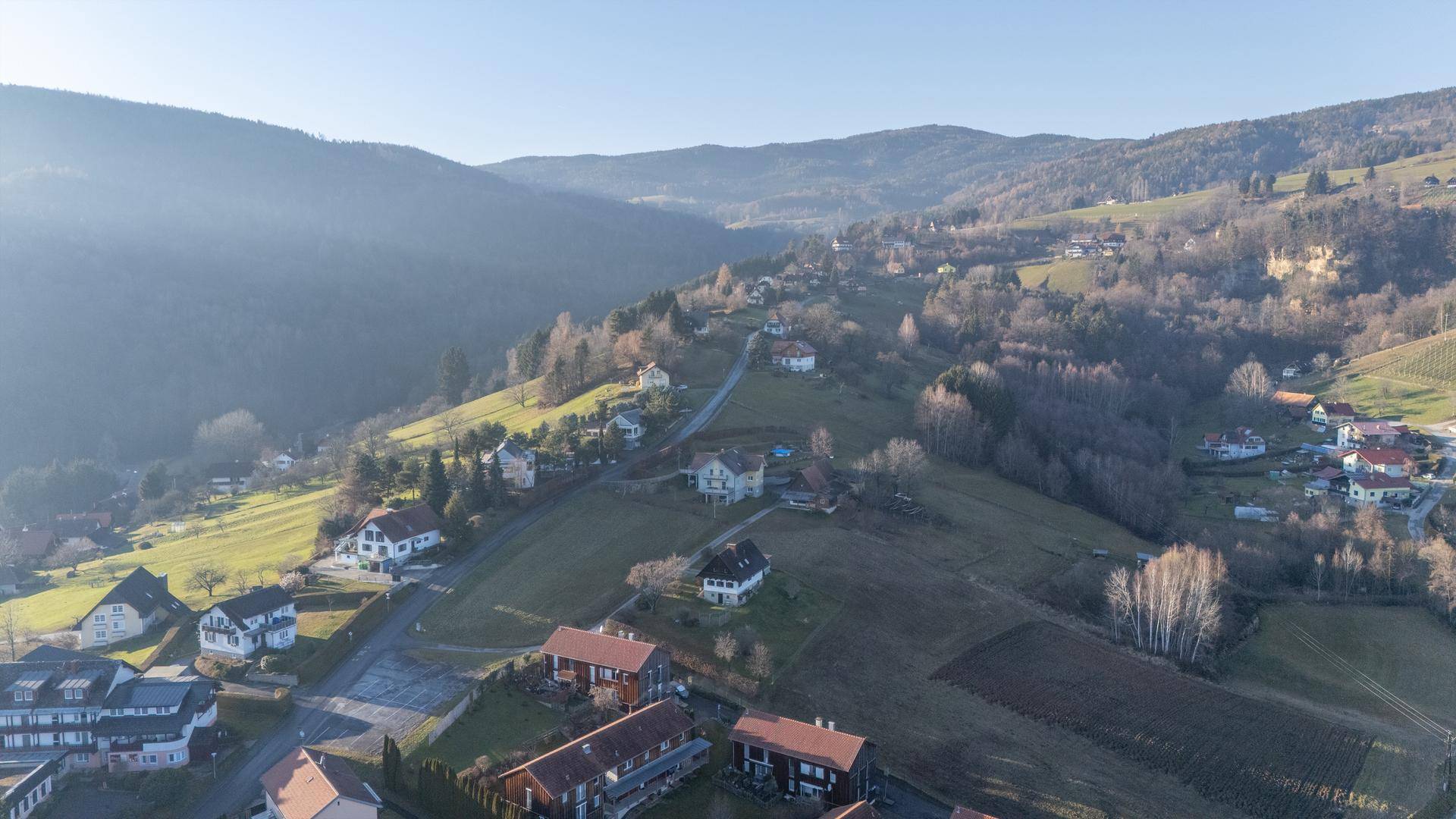 Luftaufnahme einer hügeligen Landschaft mit Wohnhäusern, Feldern und deutlich sichtbaren Weinbergen am rechten Bildrand.