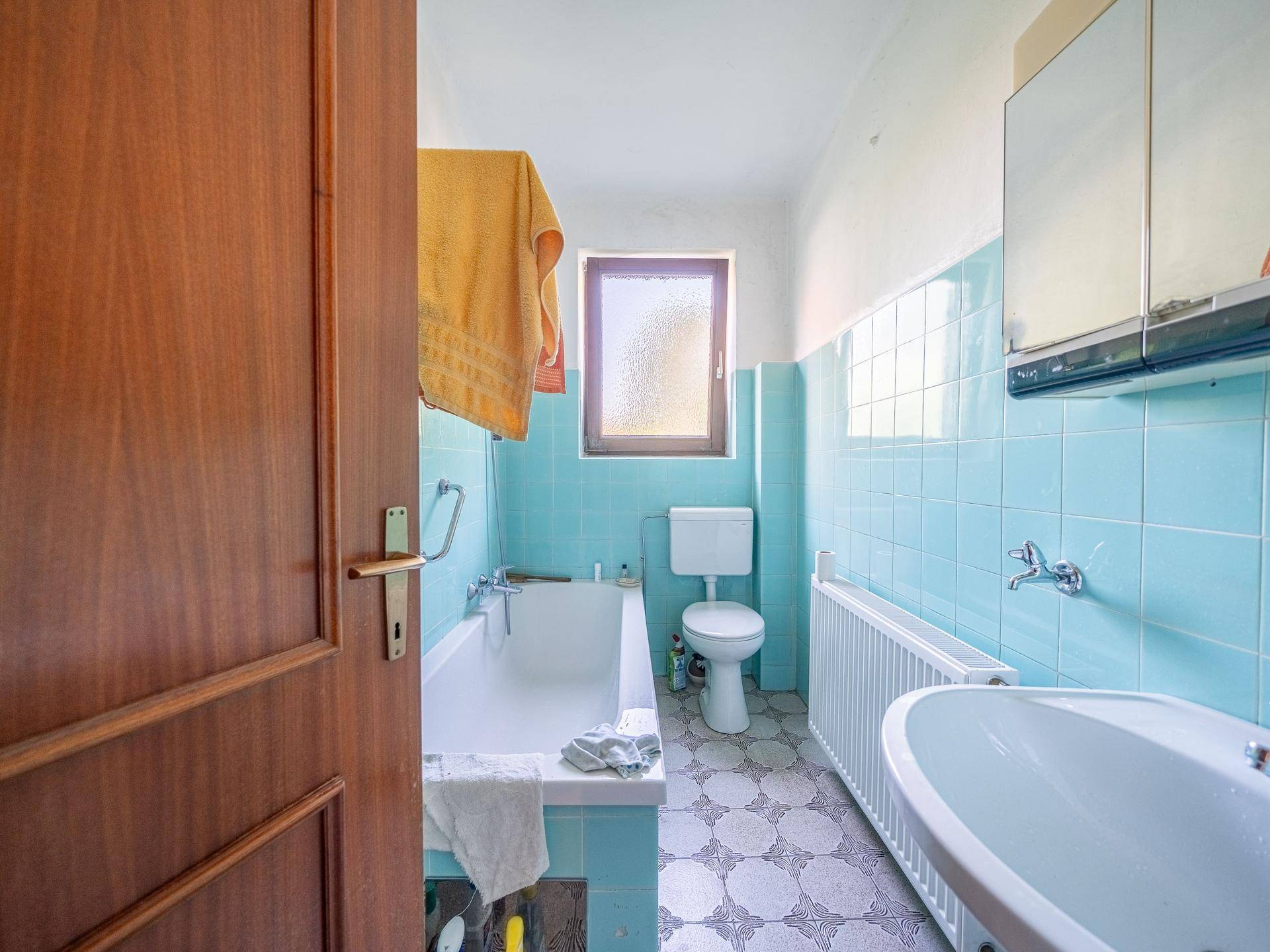 Badezimmer mit hellblauen Fliesen, Badewanne, WC und Fenster für Tageslicht.