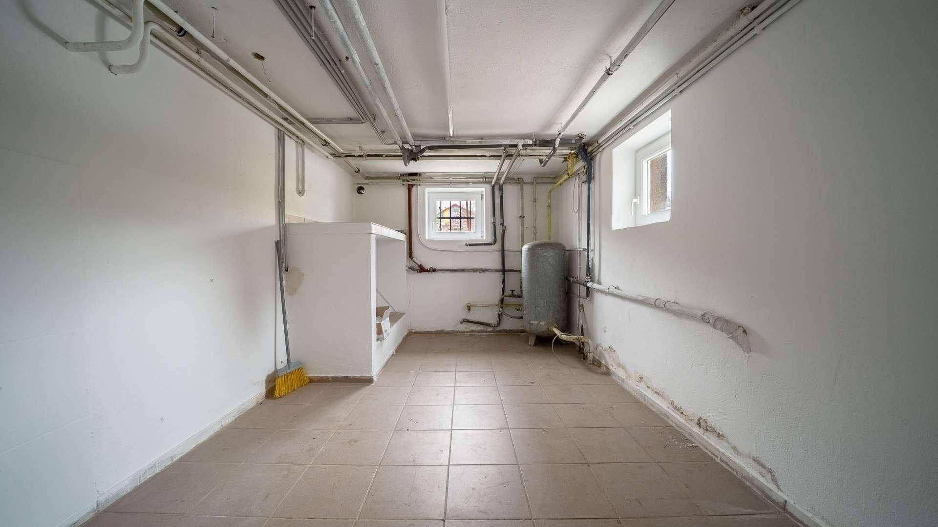 Kellerraum mit Fliesenboden, sichtbaren Rohren, Heizungsanlage und kleinem Fenster.