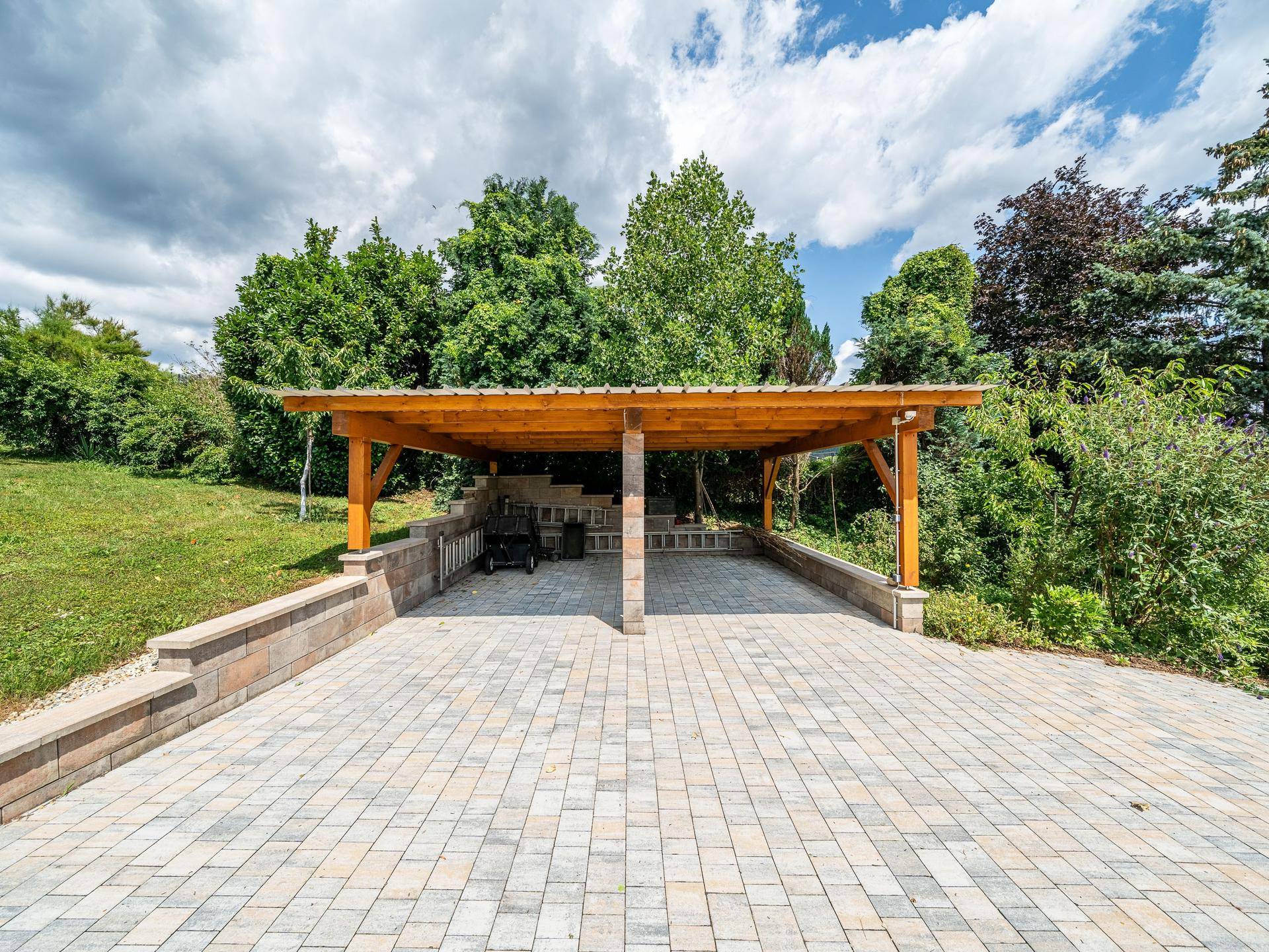 Geräumiger Carport mit Holzkonstruktion und gepflastertem Vorplatz, umgeben von Bäumen.