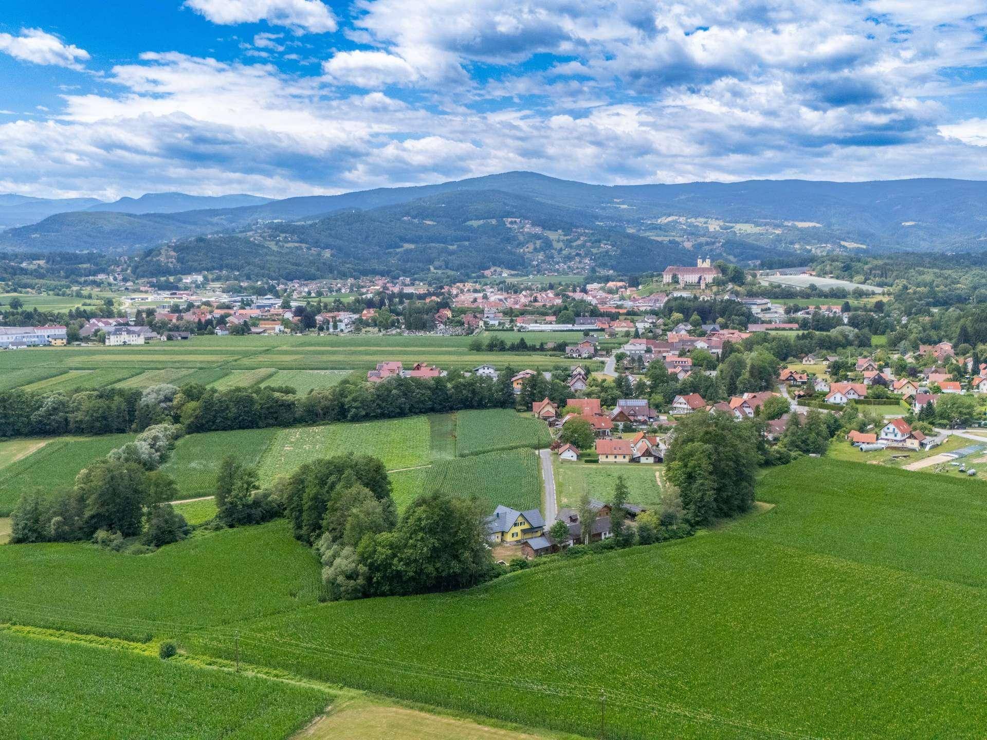 Weitläufige Luftaufnahme einer ländlichen Ortschaft mit Feldern und Bergen im Hintergrund.