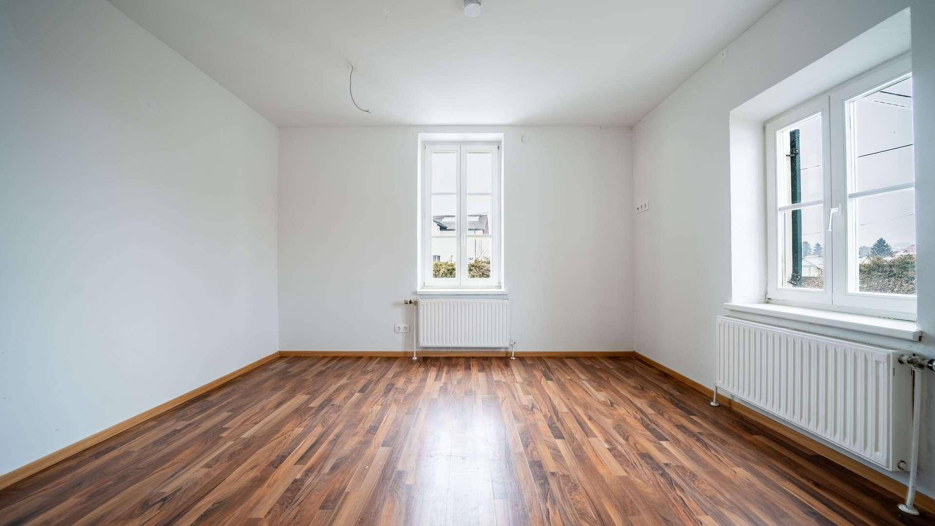 Helles Wohnzimmer mit zwei Fenstern, dunklem Laminatboden und weiß gestrichenen Wänden.