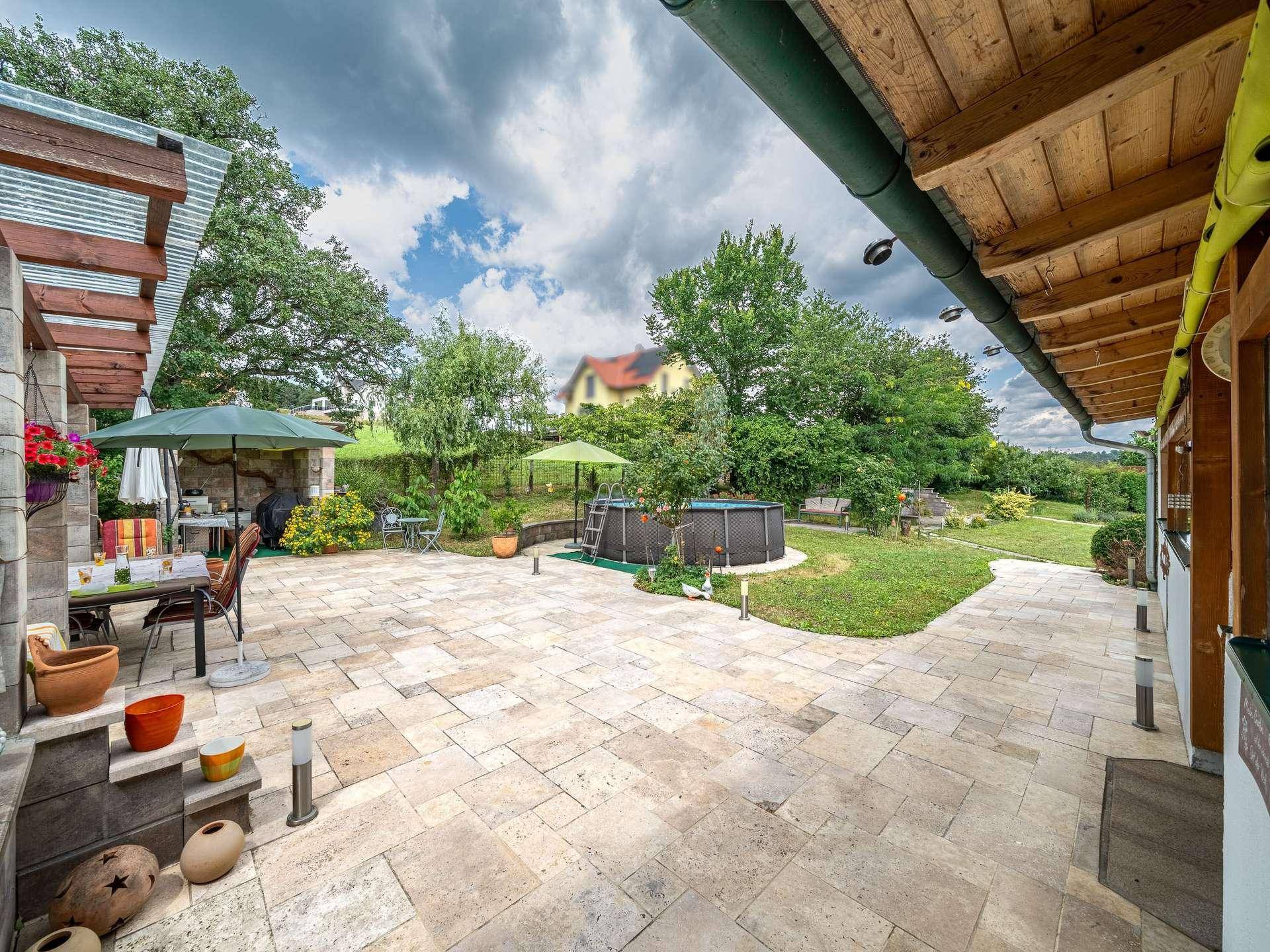 Große gepflasterte Terrasse mit Sitzgelegenheiten, die zu einem Rasenbereich mit Aufstellpool führt.