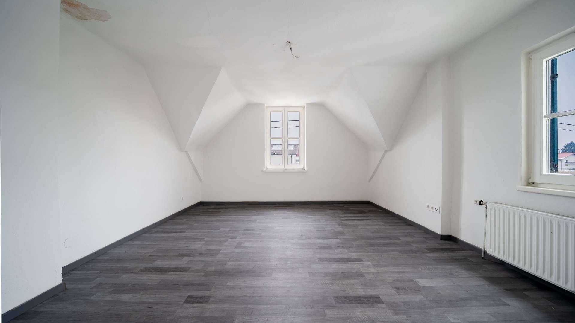 Helles Dachzimmer mit schrägen Wänden, Fenster und grauem Laminatboden.