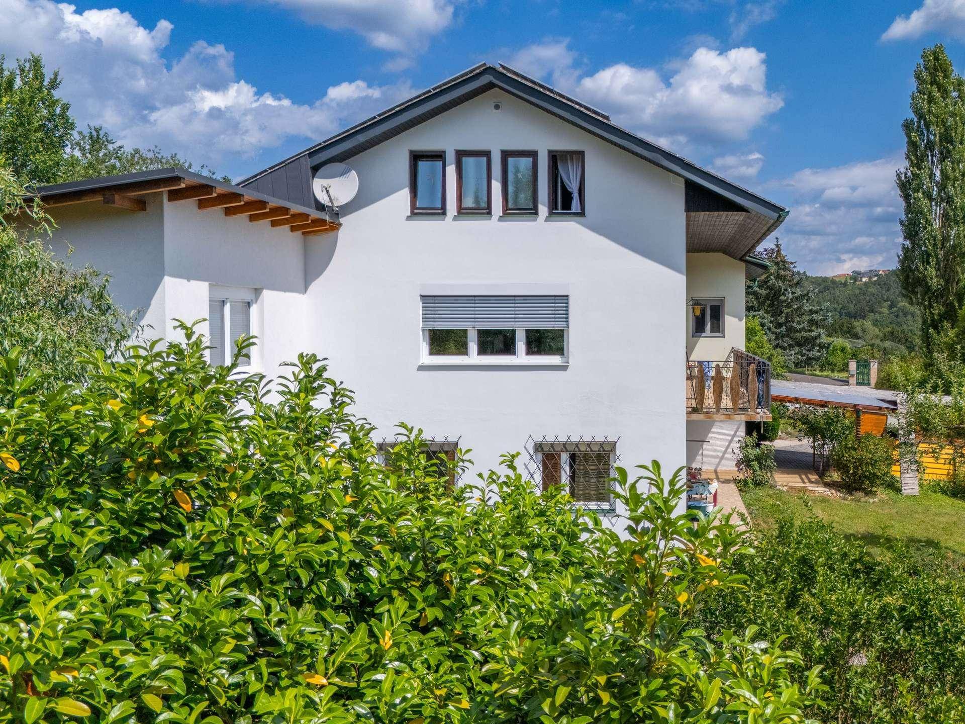 Weiße Hausfassade mit dunklem Dach und Fenstern, teilweise von grünen Büschen verdeckt.