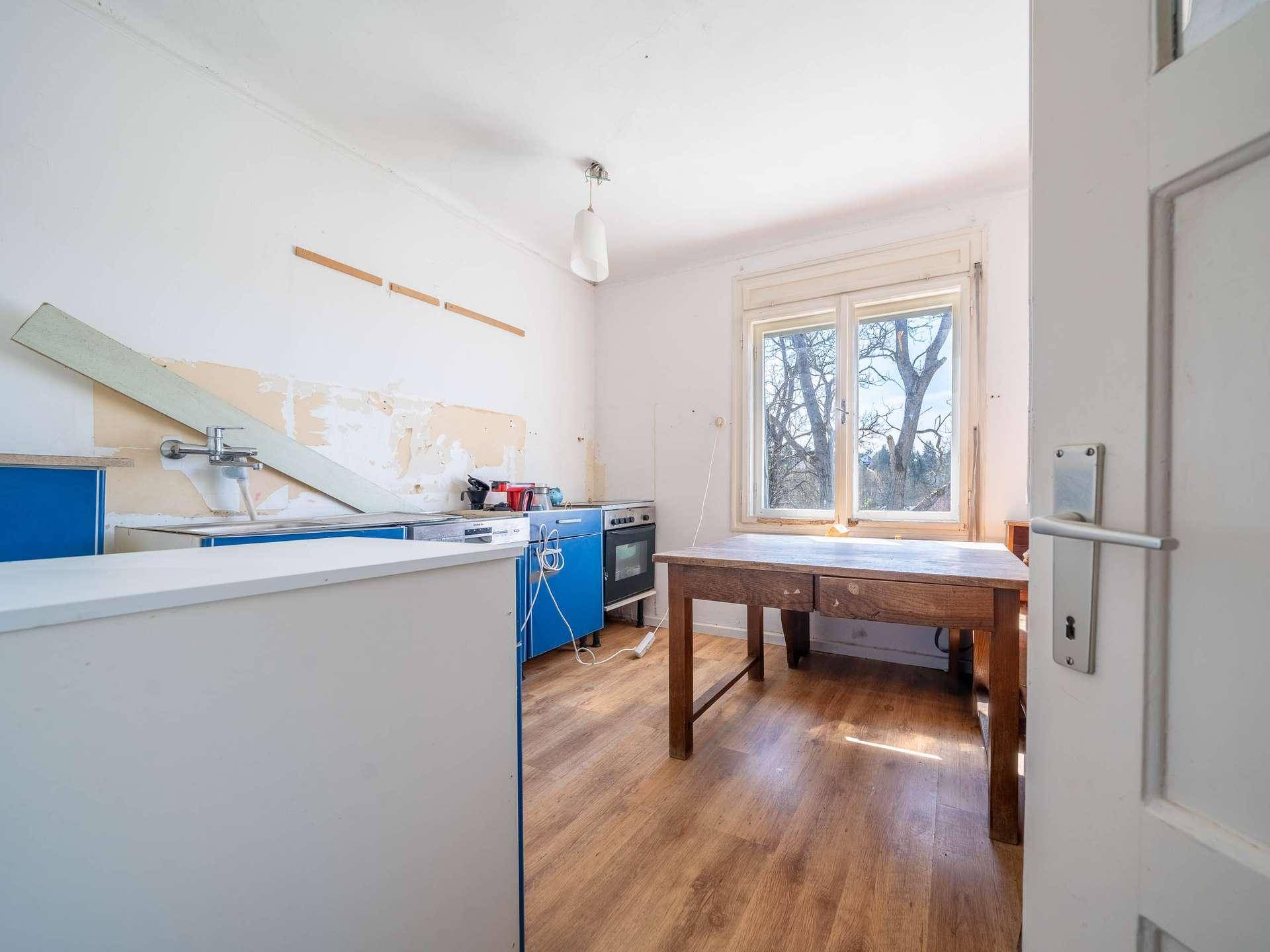 Küche mit blauen Unterschränken, Holztisch und Fenster, die eine Renovierung benötigt.