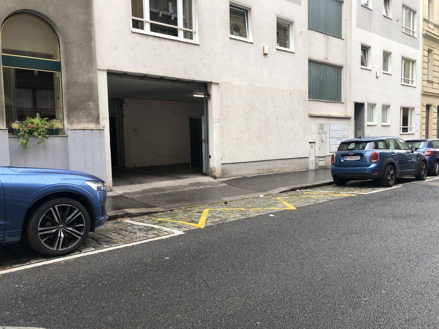 Straßenszene mit einem Garageneingang und geparkten Autos am Straßenrand vor einem Wohngebäude.