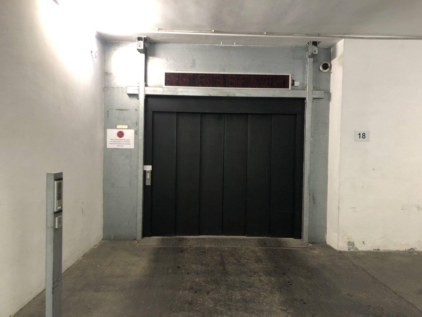 Geschlossene Aufzugstür in einer Tiefgarage, daneben eine Gegensprechanlage und eine Überwachungskamera.