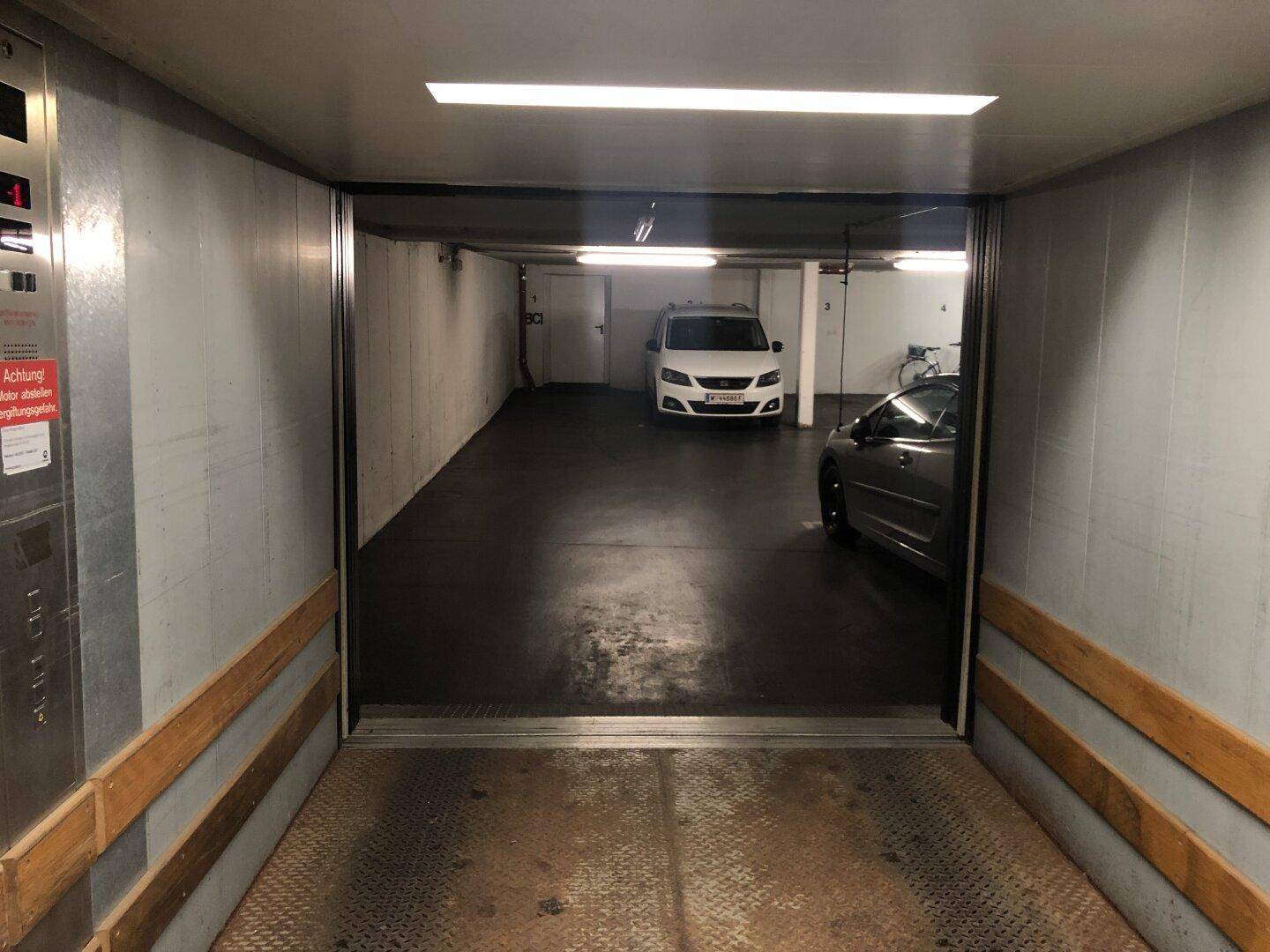 Blick aus dem geöffneten Lastenaufzug in die Tiefgarage mit geparkten Autos und heller Beleuchtung.