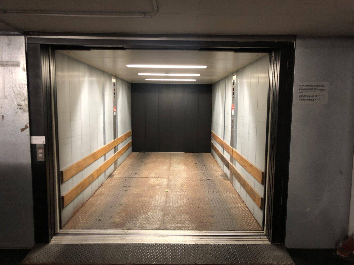 Großer Lastenaufzug mit geöffneten Türen, geeignet für Fahrzeuge, ausgestattet mit Schutzleisten und Beleuchtung.