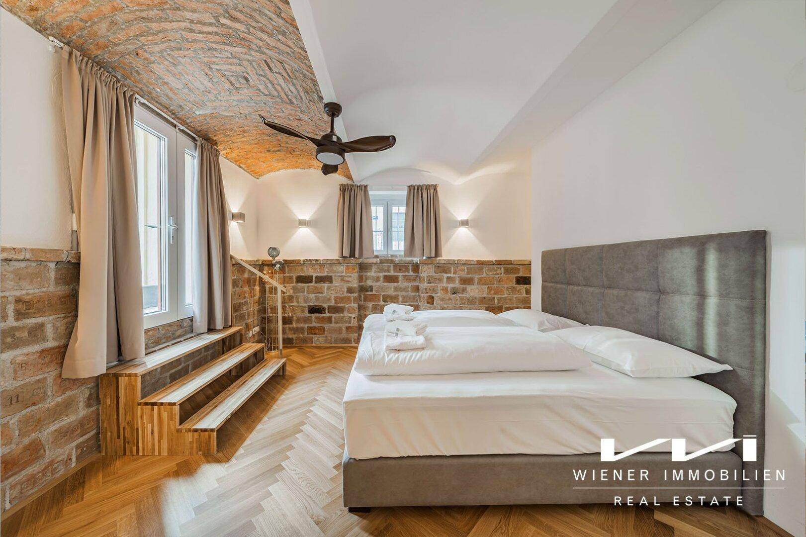 Schlafzimmer mit großem Bett, freigelegten Ziegelwänden, Fischgrätparkett und einer beeindruckenden Gewölbedecke.