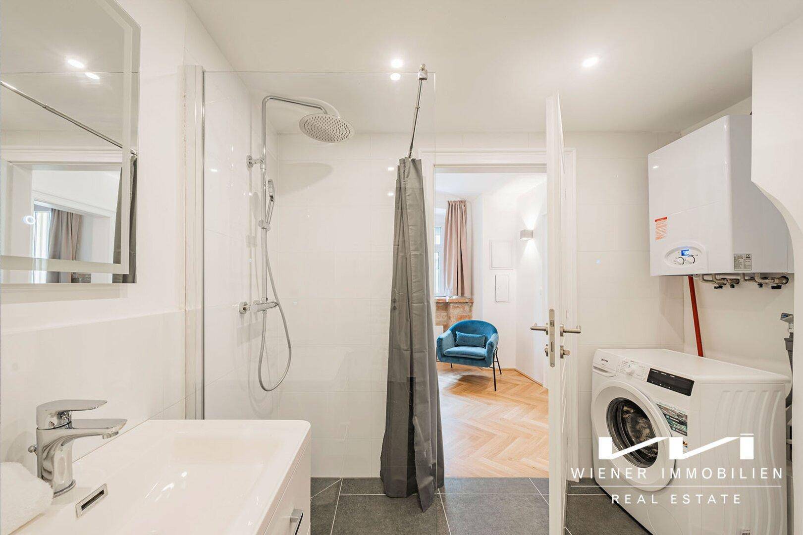 Modernes Badezimmer mit Duschkabine, Waschbecken und Waschmaschine, ausgestattet mit weißen Fliesen und grauen Bodenfliesen.