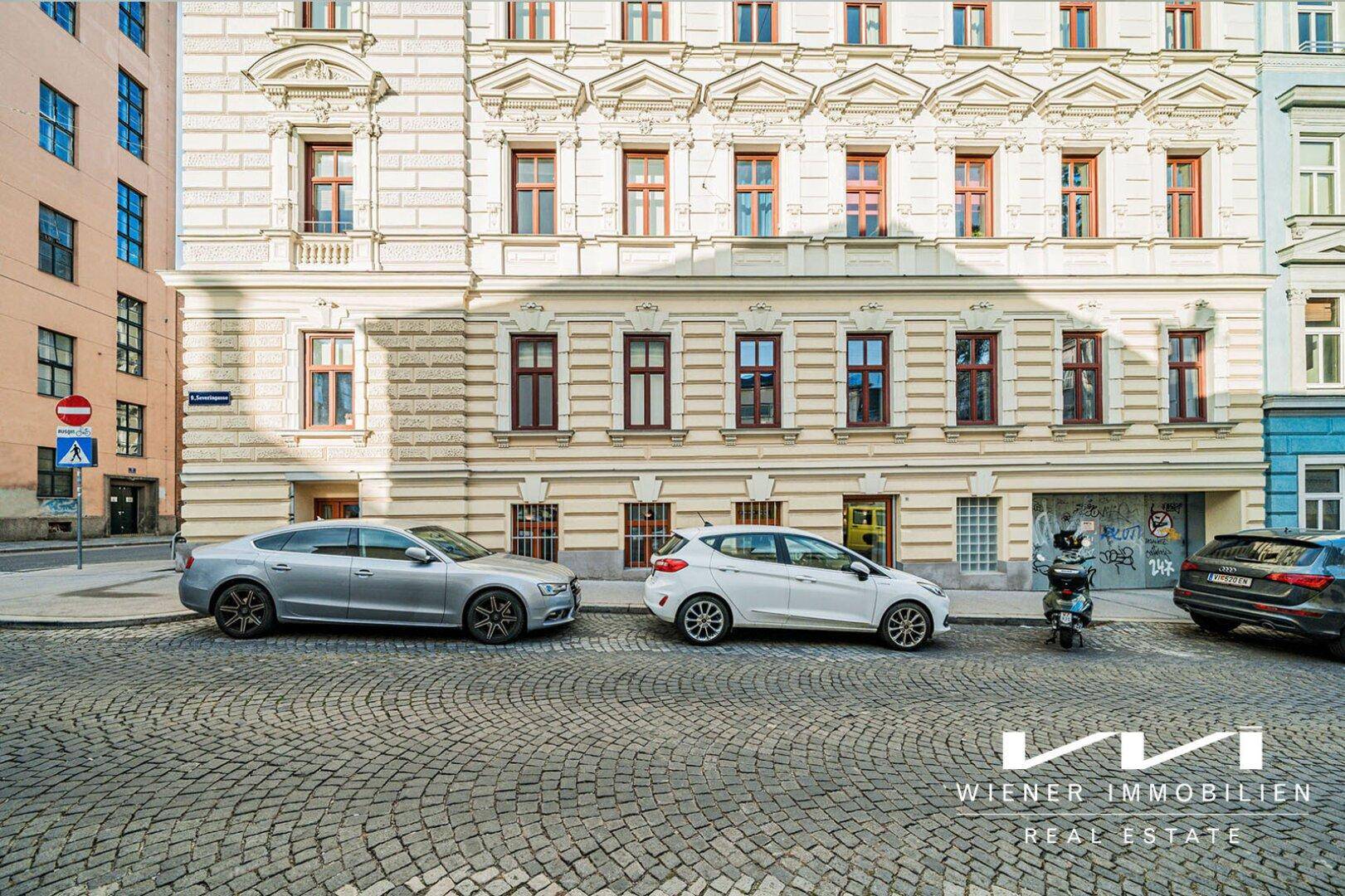 Historische Gebäudefassade mit kunstvollen Details und geparkten Autos auf einer Kopfsteinpflasterstraße.