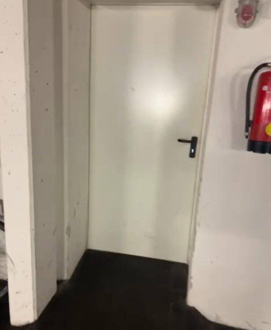 Eine schlichte weiße Tür in einem Keller- oder Technikbereich, daneben ein Feuerlöscher an der Wand.