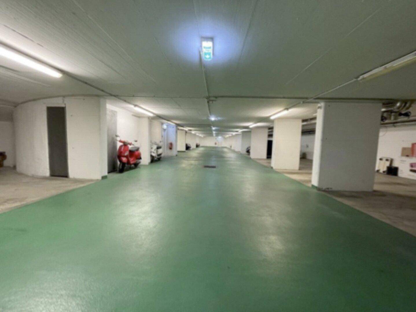 Geräumige Tiefgarage mit grünem Boden, weißen Säulen und Deckenbeleuchtung, bietet Stellplätze für Fahrzeuge.