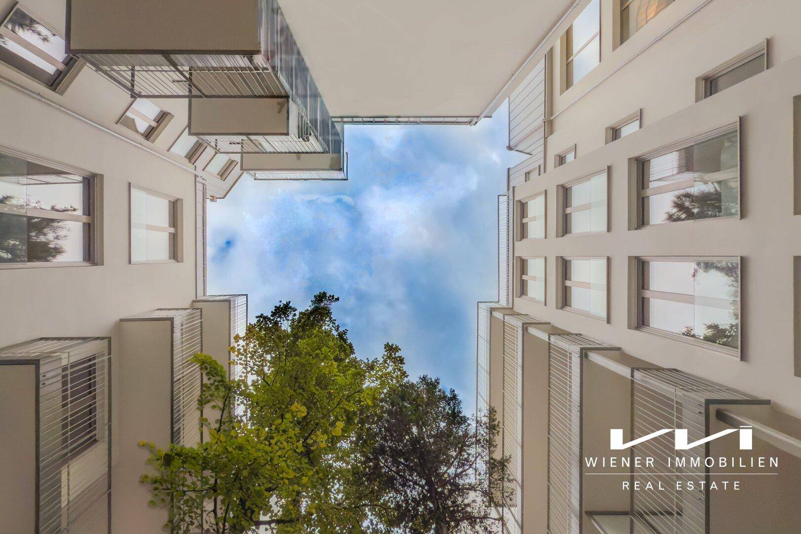 Blick nach oben in einen hellen Innenhof, umgeben von modernen Gebäudefassaden und Bäumen.