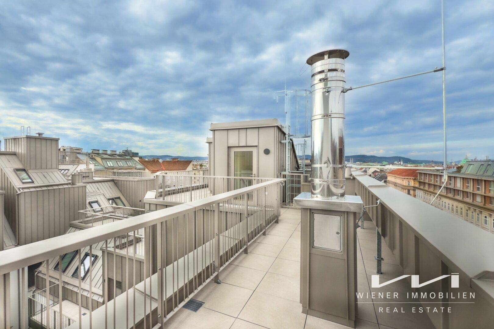 Geräumige Dachterrasse mit Fliesenboden, Geländer und weitem Blick über die Stadt.