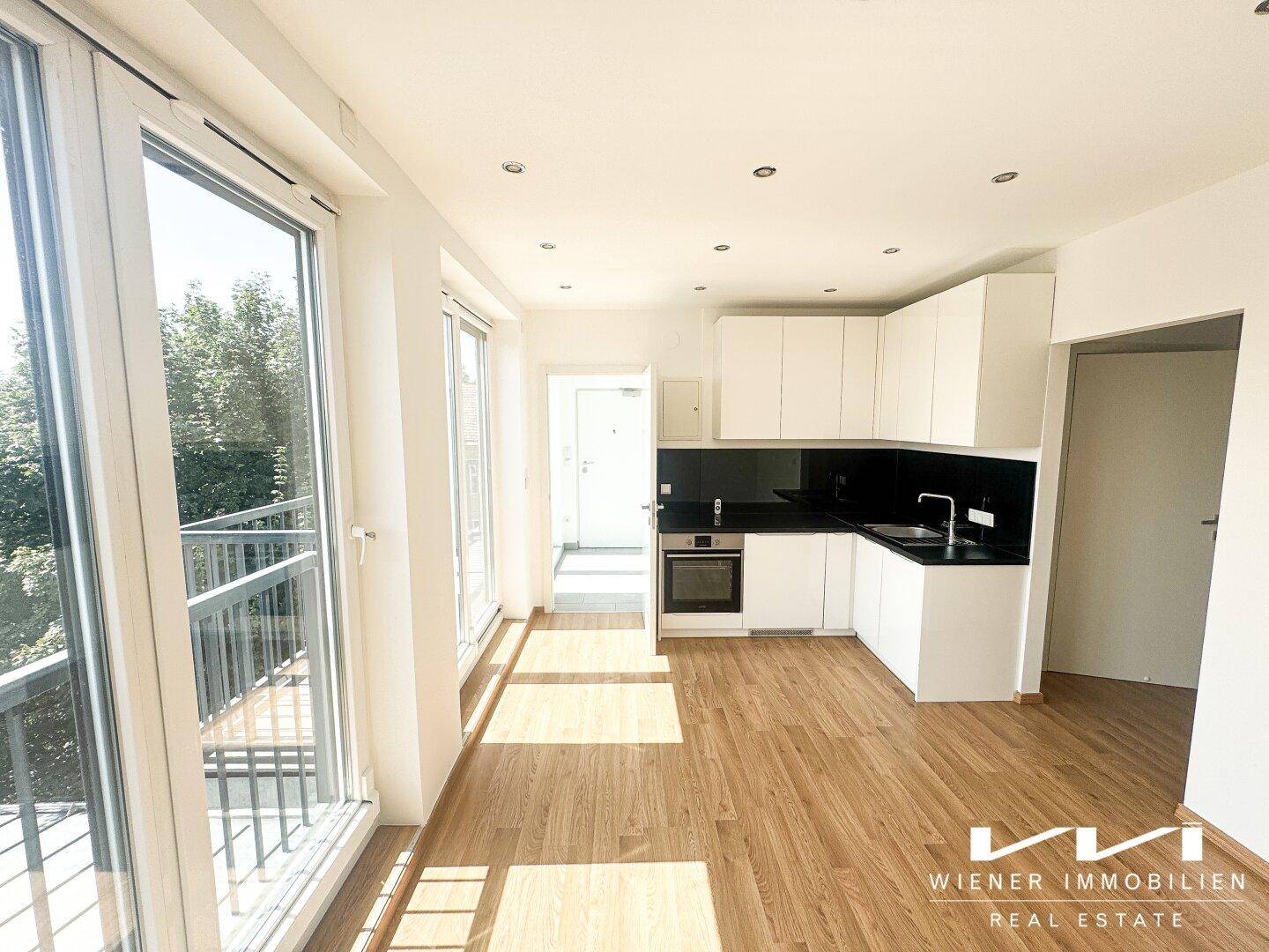 Heller offener Wohnbereich mit integrierter Küche, Holzboden und Zugang zum Balkon.