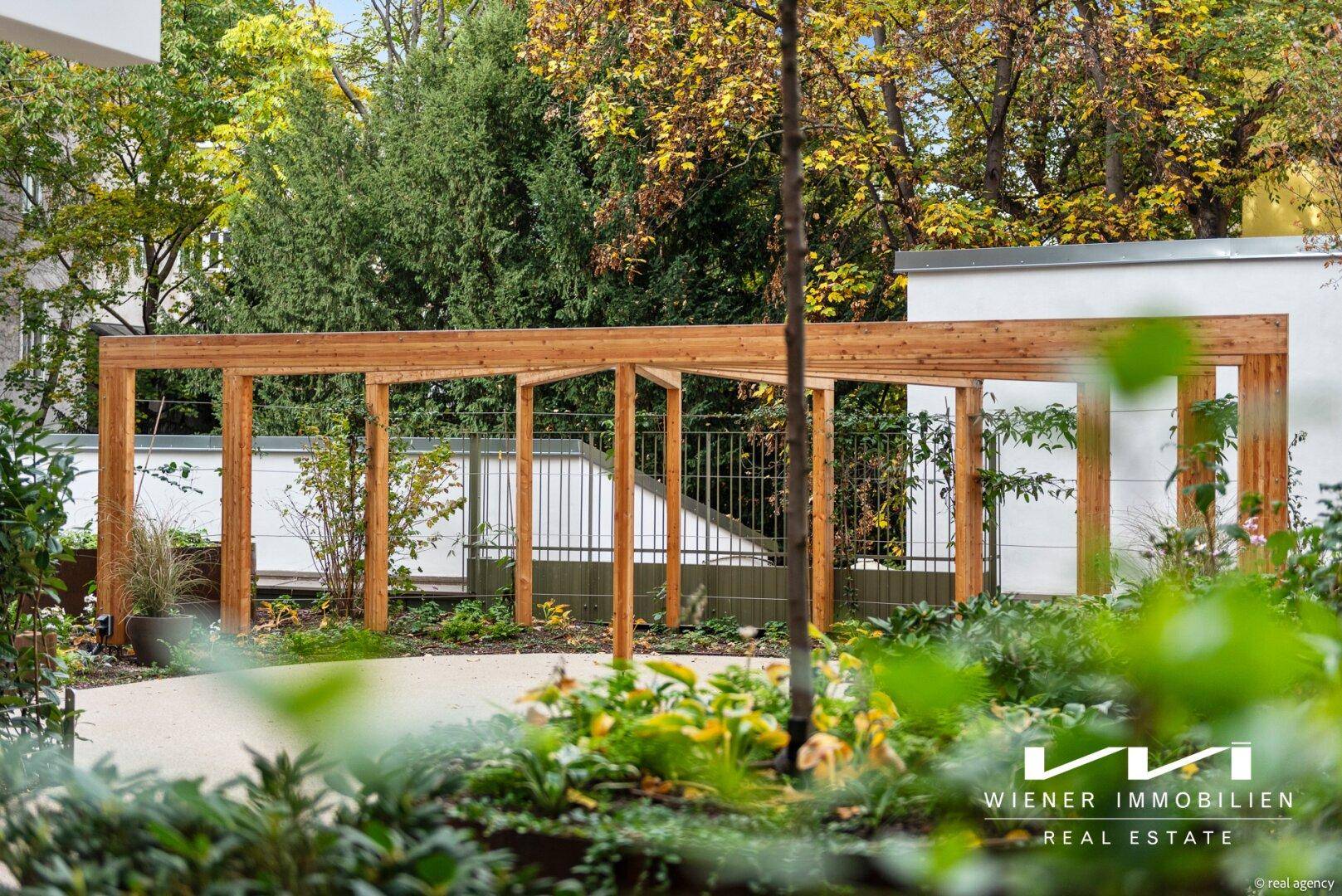 Holzpergola in einem grünen Gartenbereich, umgeben von Bäumen und Sträuchern.