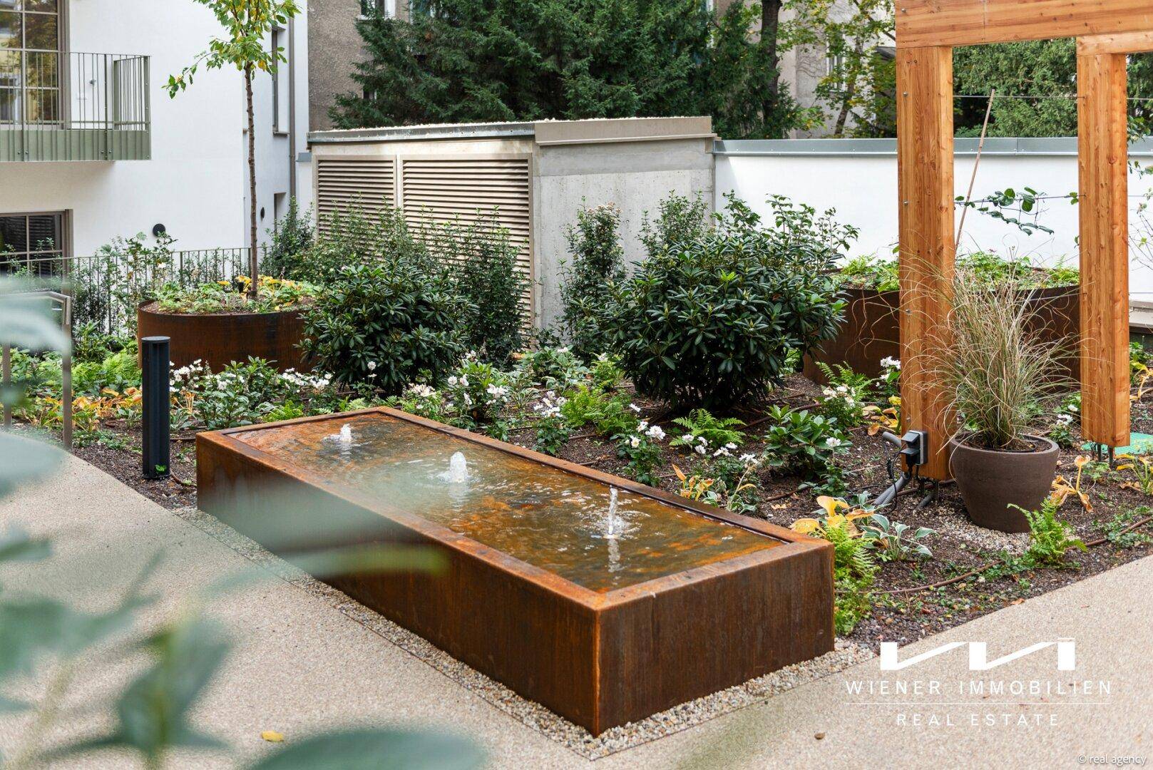 Detailansicht eines modernen Gemeinschaftsgartens mit Wasserbecken, Pflanzen und Holzpergola.