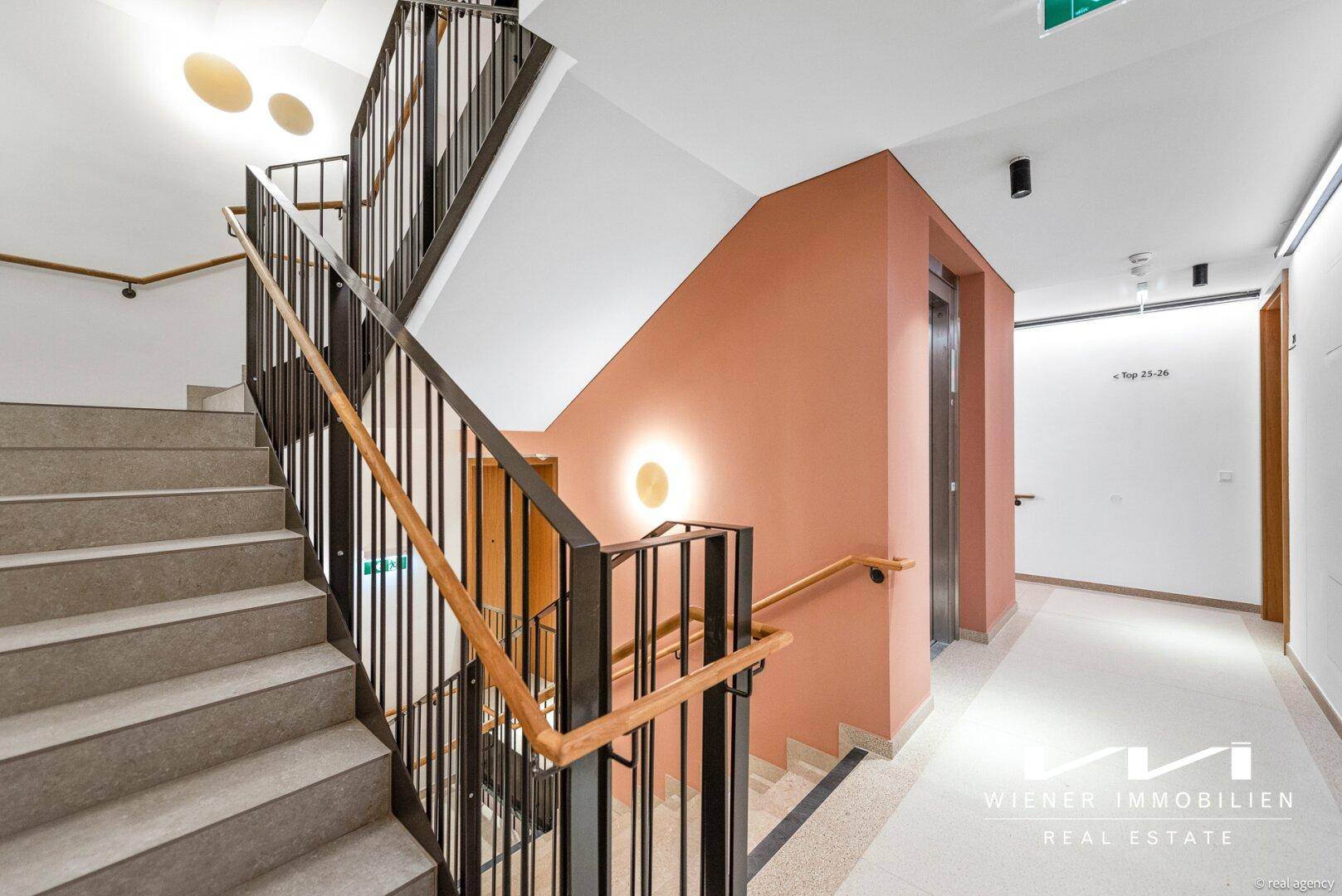 Modernes Treppenhaus mit Aufzug, dunklem Geländer und farbiger Wandgestaltung.