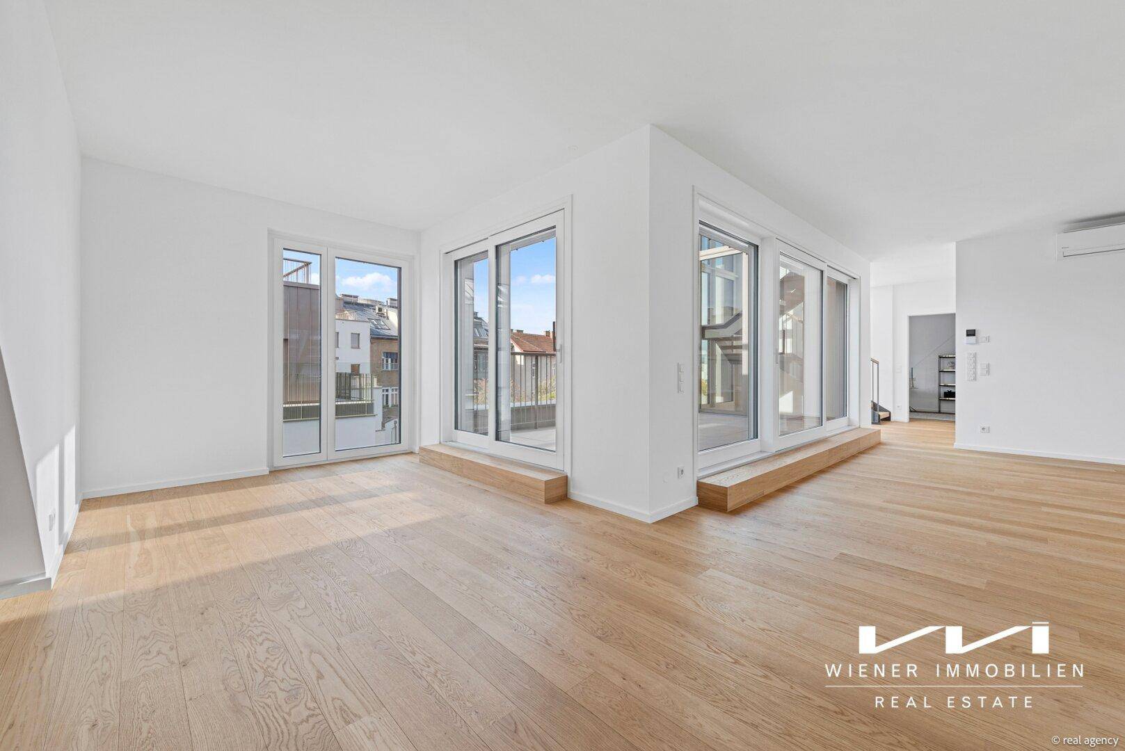 Geräumiger, leerer Wohnraum mit hellem Holzboden und großen Fenstern, die viel Licht hereinlassen.