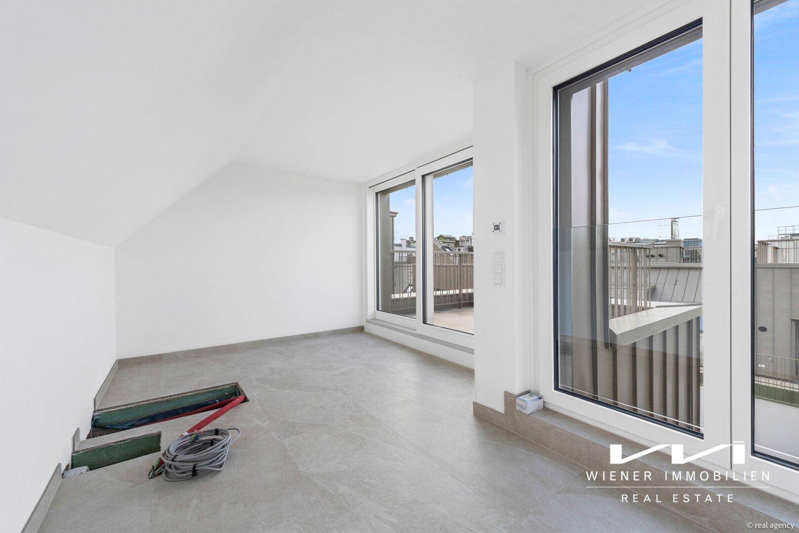 Raum im Bauzustand mit großen Fenstern und Blick auf die Dächer der Stadt.