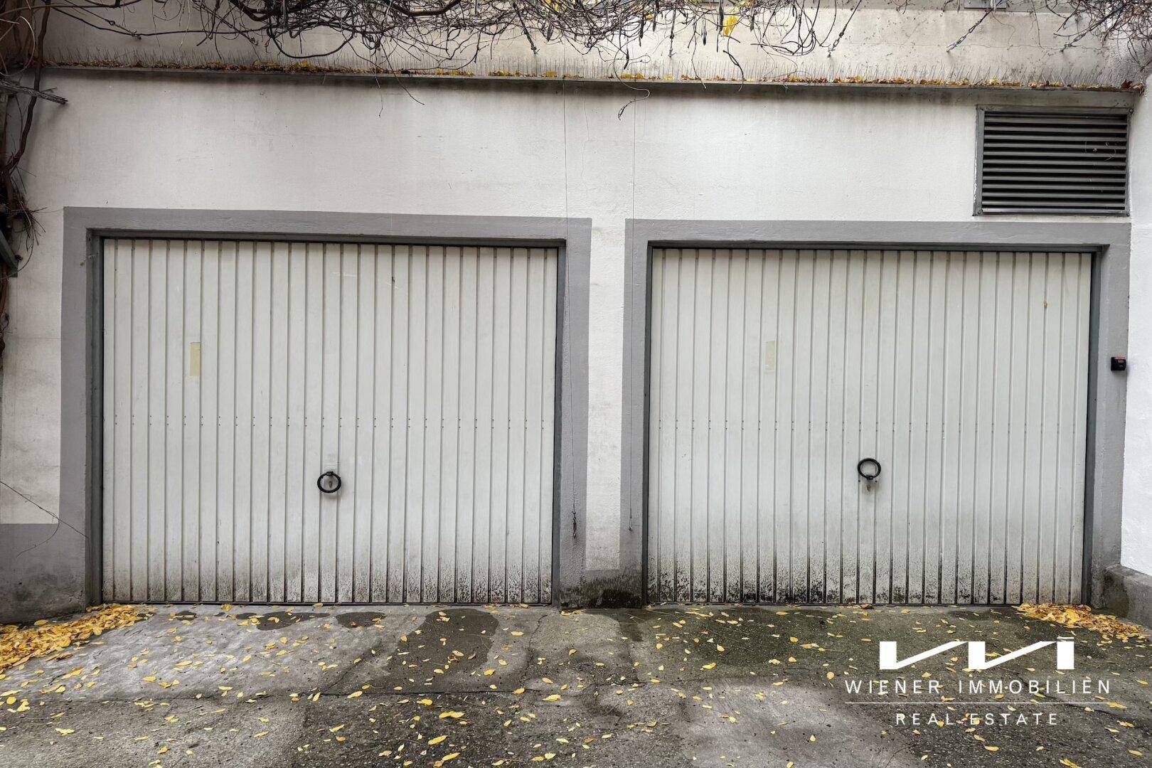 Zwei weiße Garagentore mit grauen Rahmen und einer Betonwand darüber, umgeben von trockenem Laub.