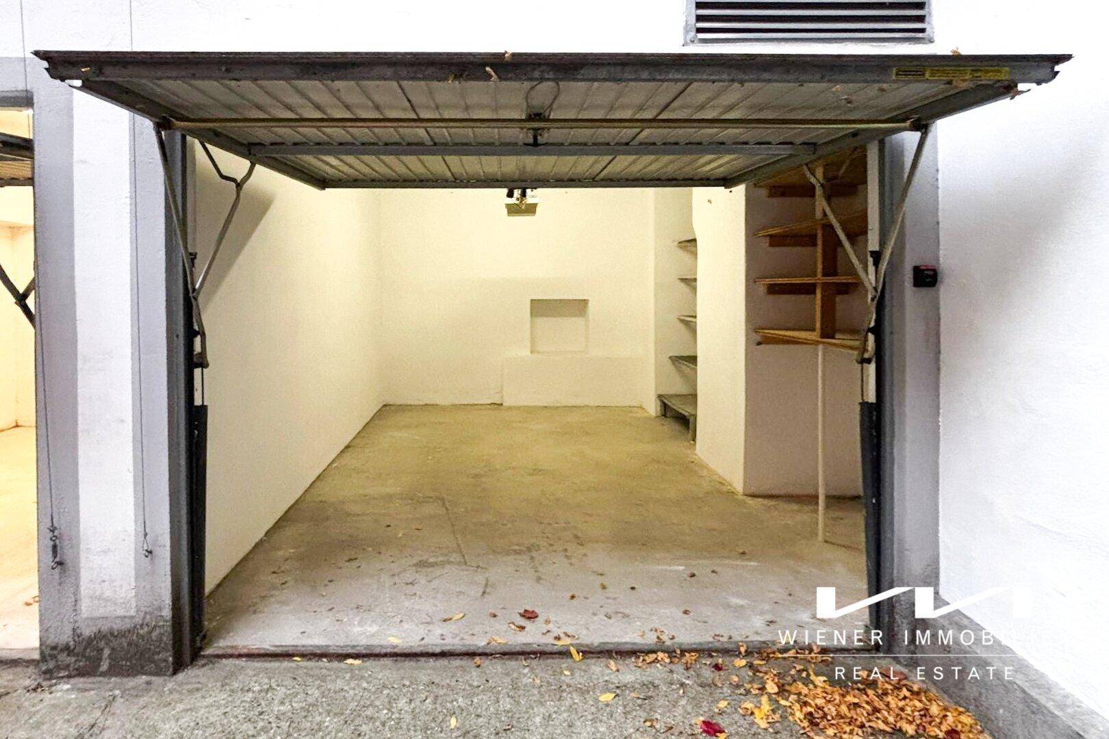 Innenansicht einer Garage mit weißen Wänden, Betonboden und einfachen Holzregalen an der rechten Seite.